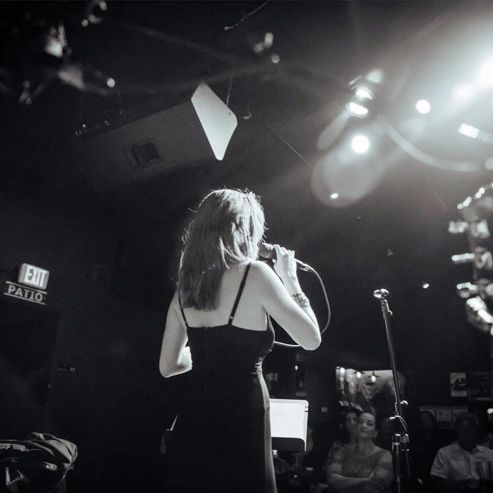 Woman singing on stage with microphone, audience in background, dim lighting, black dress, hair down.