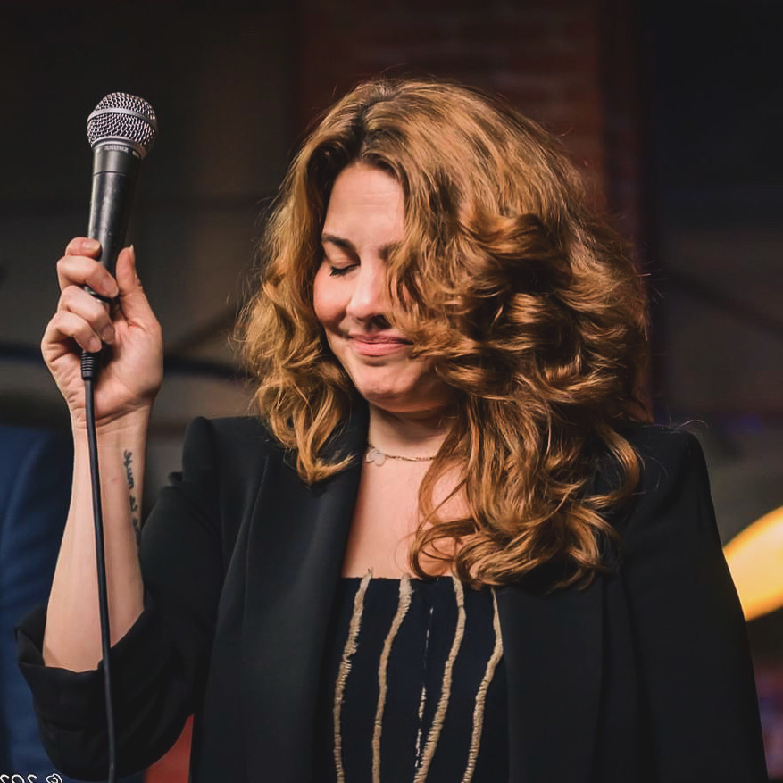 Woman with curly hair holding a microphone, smiling with eyes closed, in a dimly lit setting.