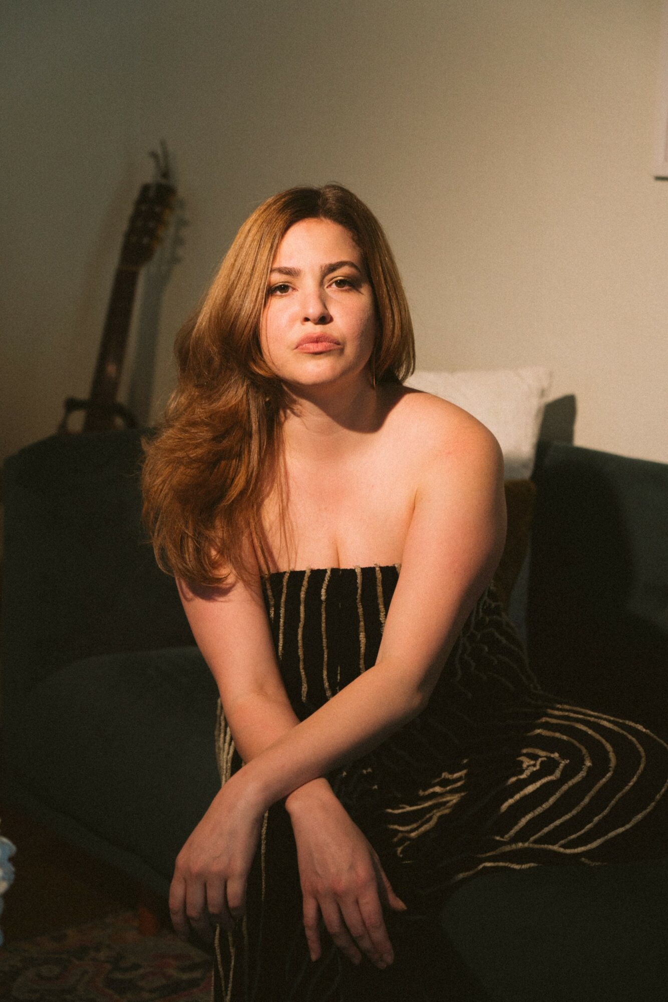Young woman with long wavy hair sitting on a dark sofa, wearing a strapless striped dress, in a dimly lit room.