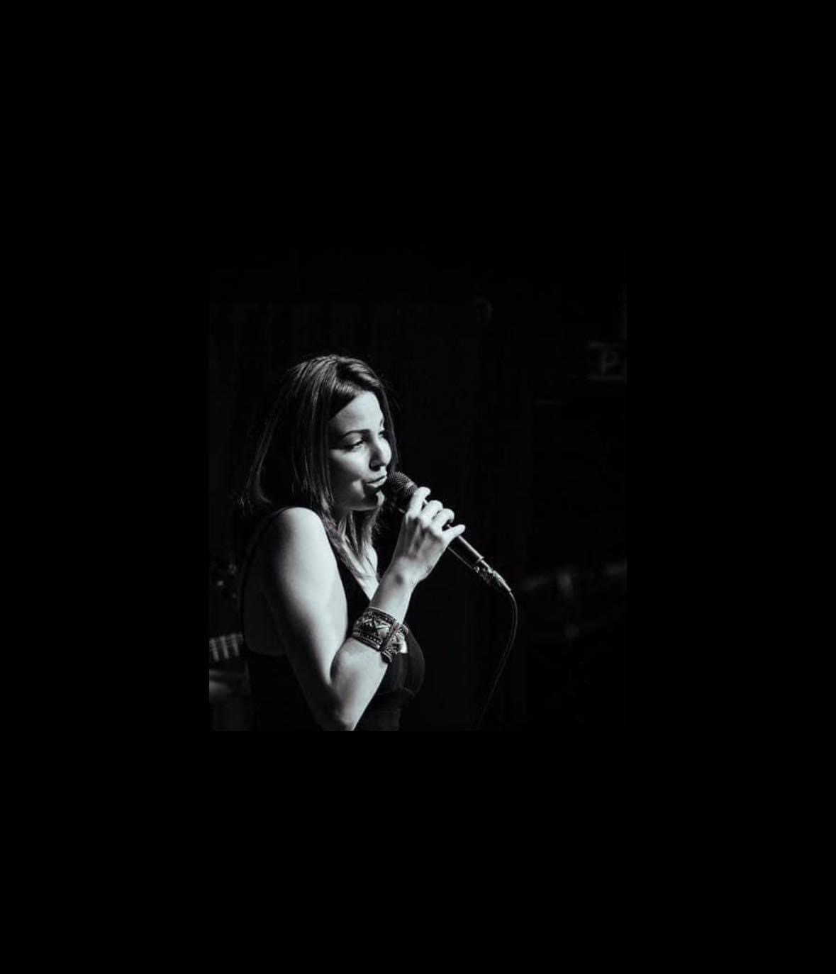 Woman singing into a microphone on stage, with dark background, in black and white.