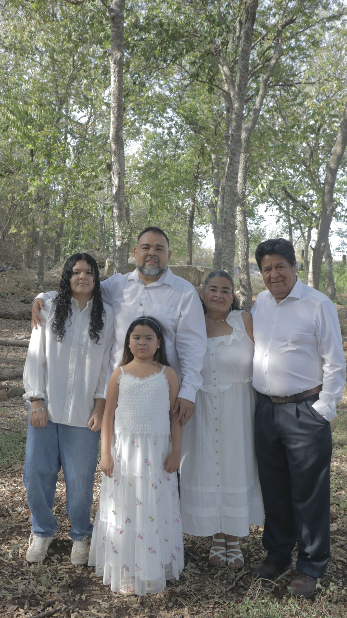 Group of five people standing outdoors among trees, some smiling, in casual and semi-formal clothing.