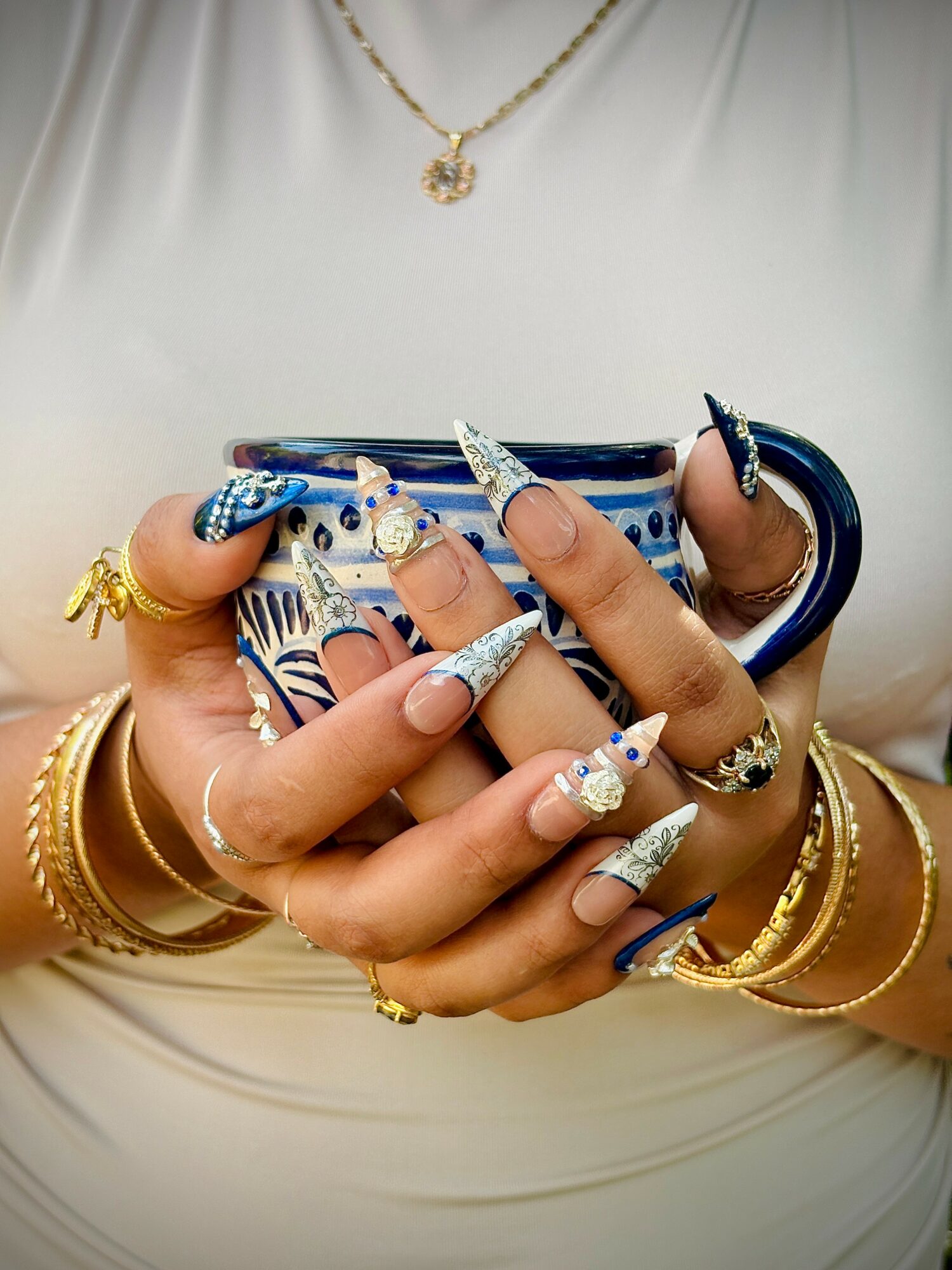 Person holding a blue and white patterned cup with decorated nails and gold jewelry, including rings and bracelets.