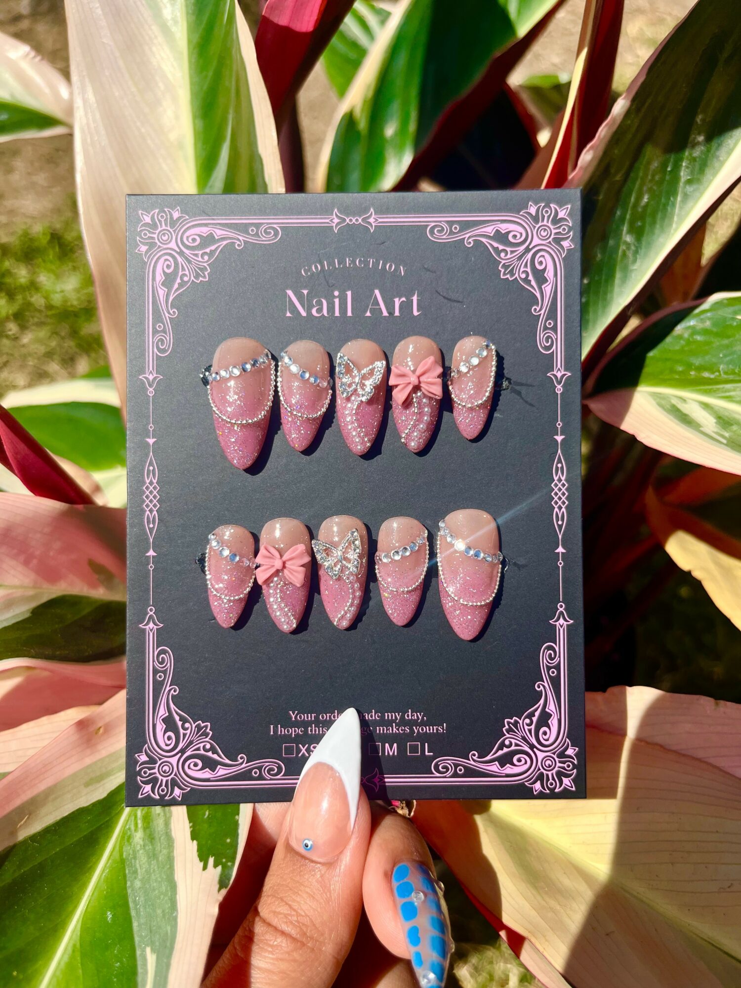 Display of pink and white nail art with rhinestones on a decorative card, held by a hand with a blue polka dot nail, surrounded by green and red leaves.