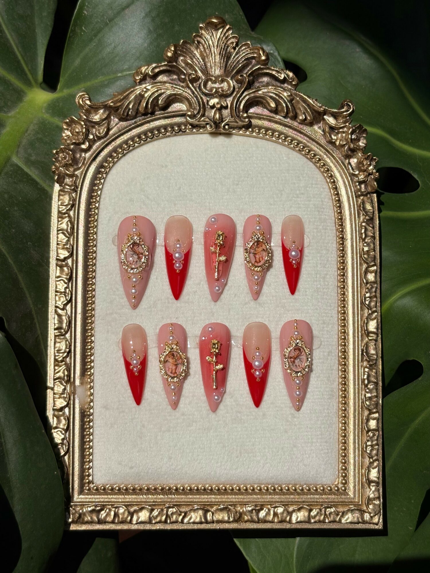 Decorative frame with ten nail art designs featuring red and nude colors with gold embellishments, set against green leaves background.