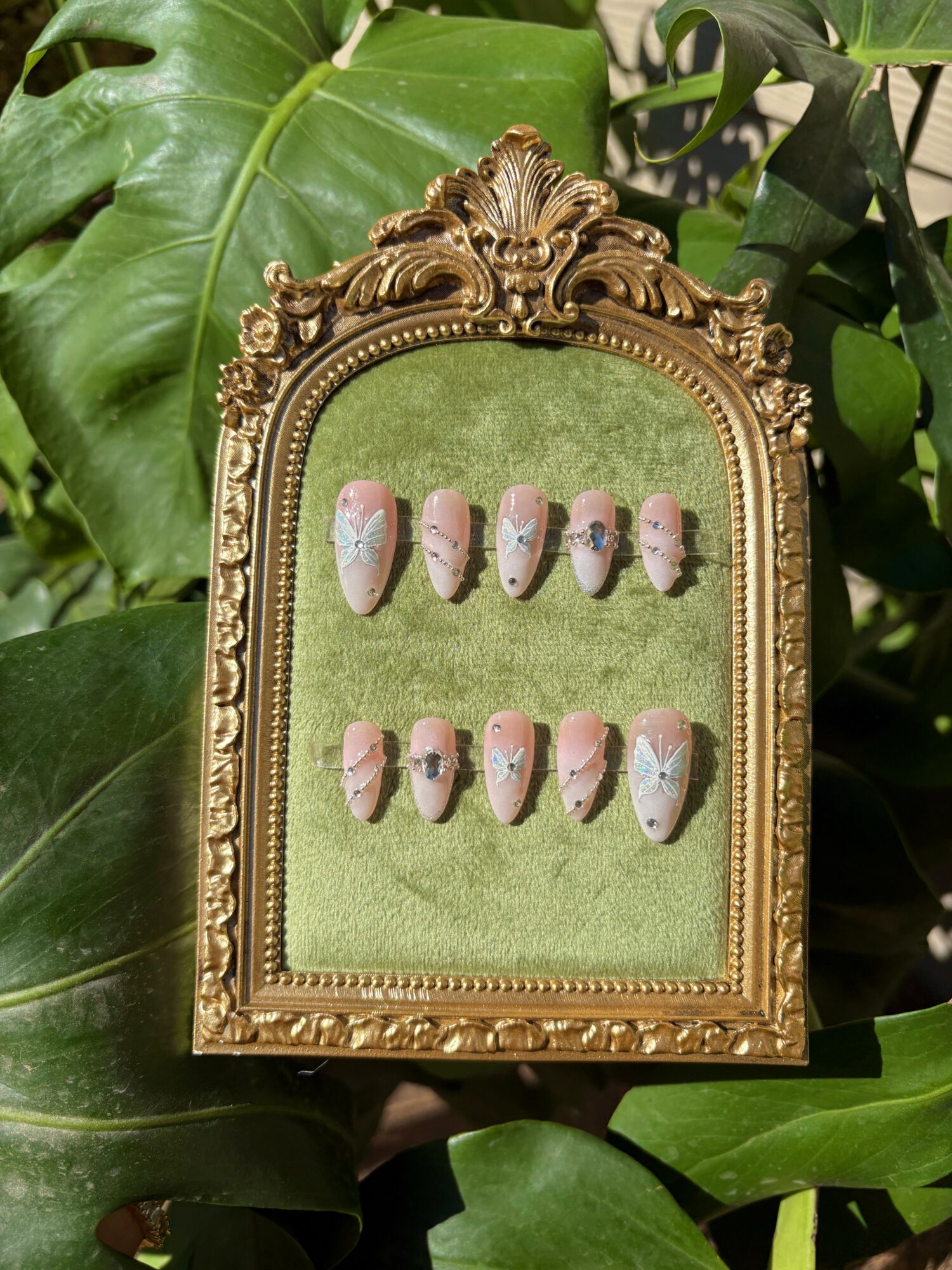 Ornate gold frame with pink and white decorated nails on green background, surrounded by large green leaves.