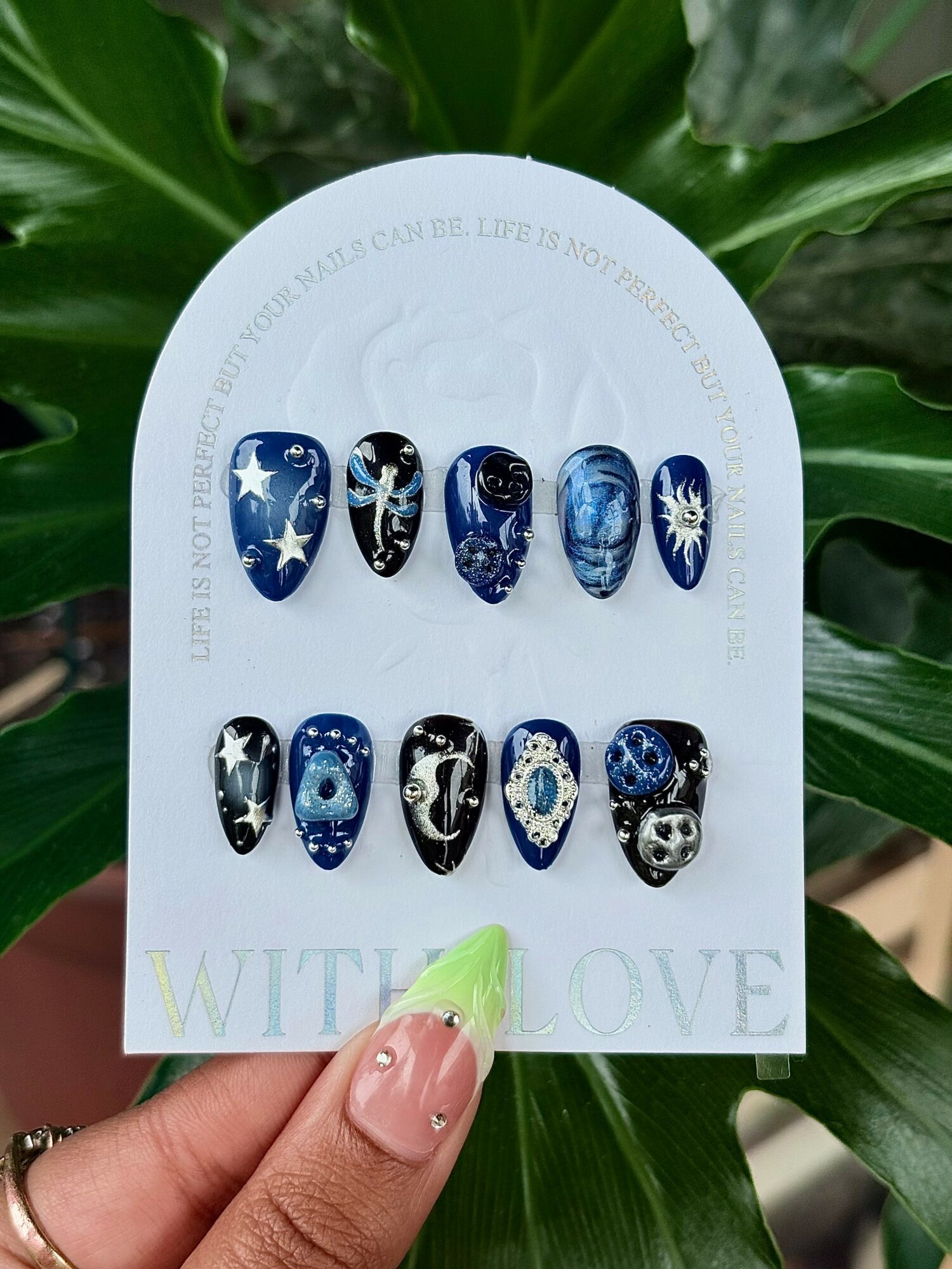 Display of 12 galaxy-themed nail art designs on a white card, held by a person with a green nail, against green leaves background.