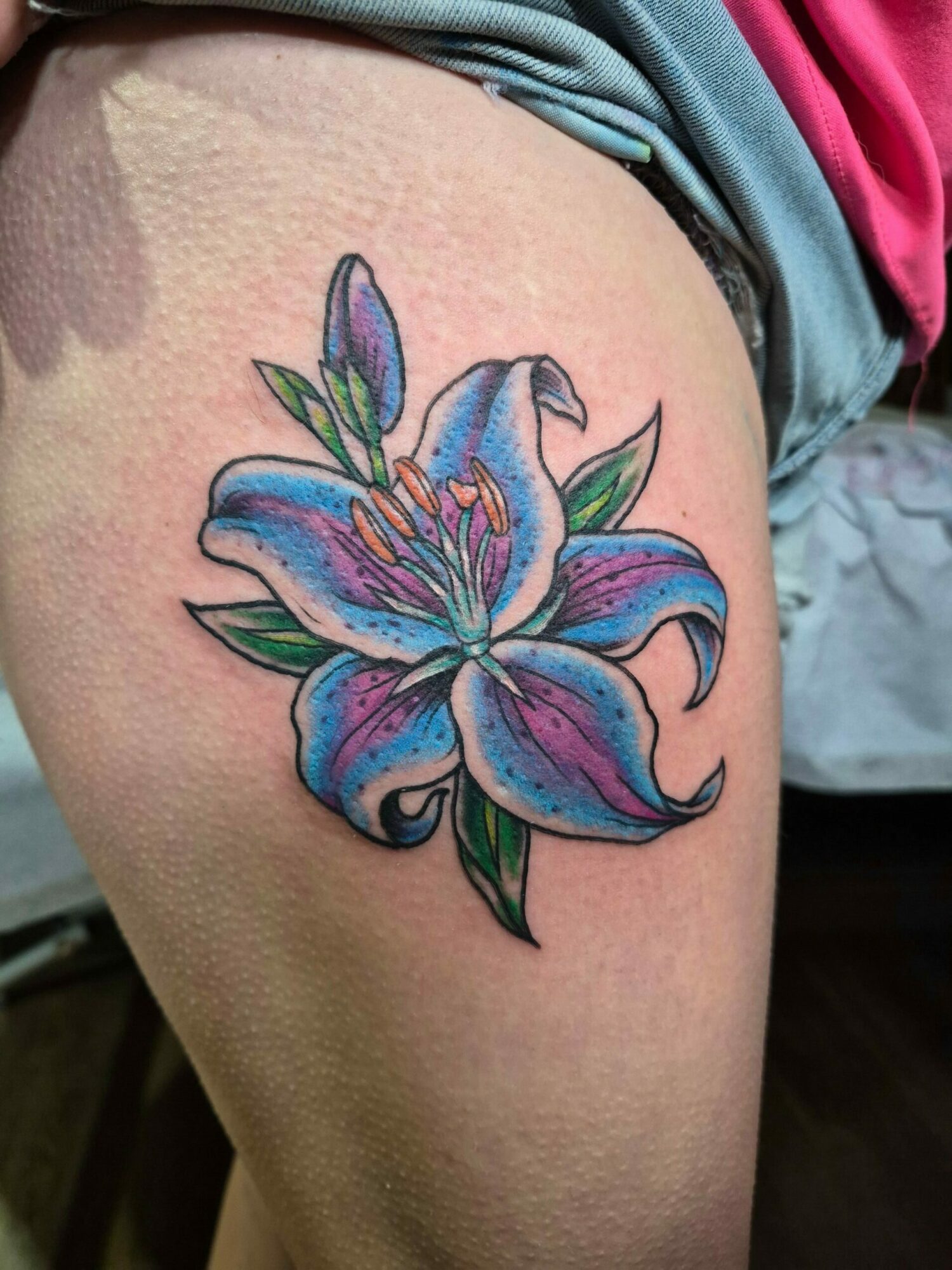 Colorful lily flower tattoo on thigh with blue, purple, and green hues, detailed petals and leaves, on light skin.