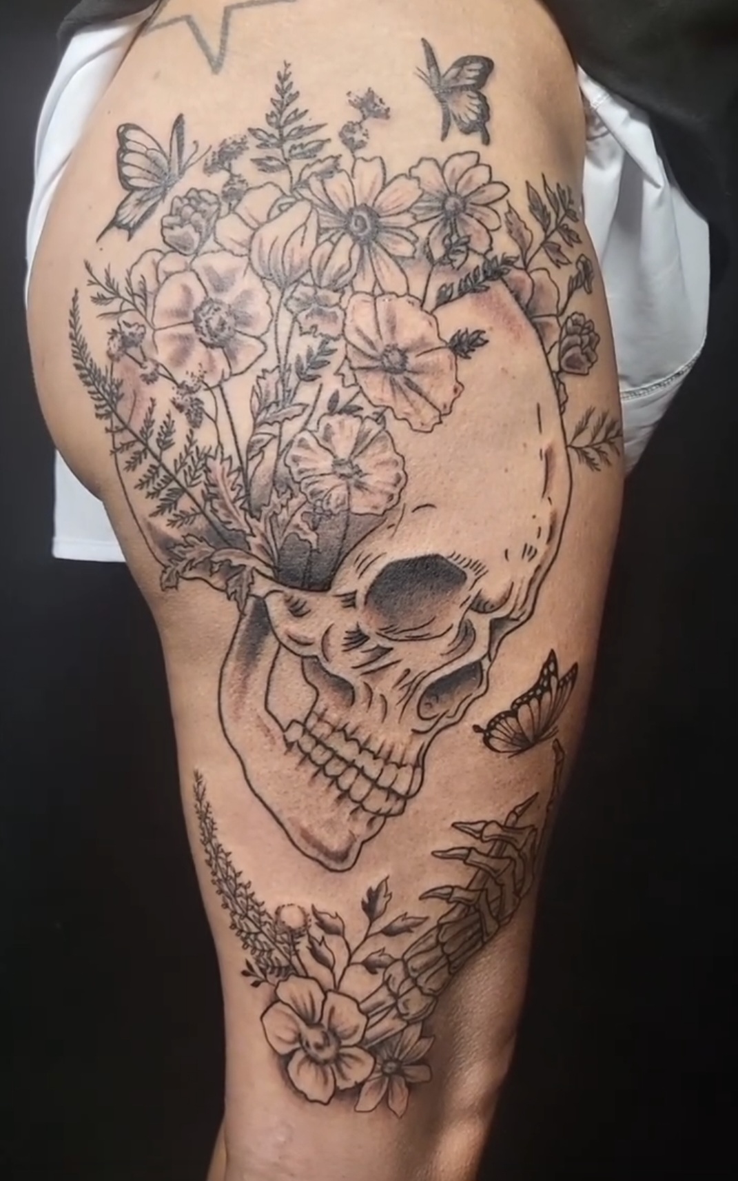 Detailed skull tattoo with flowers and butterflies on thigh, black ink design, realistic style, floral elements surrounding skull.