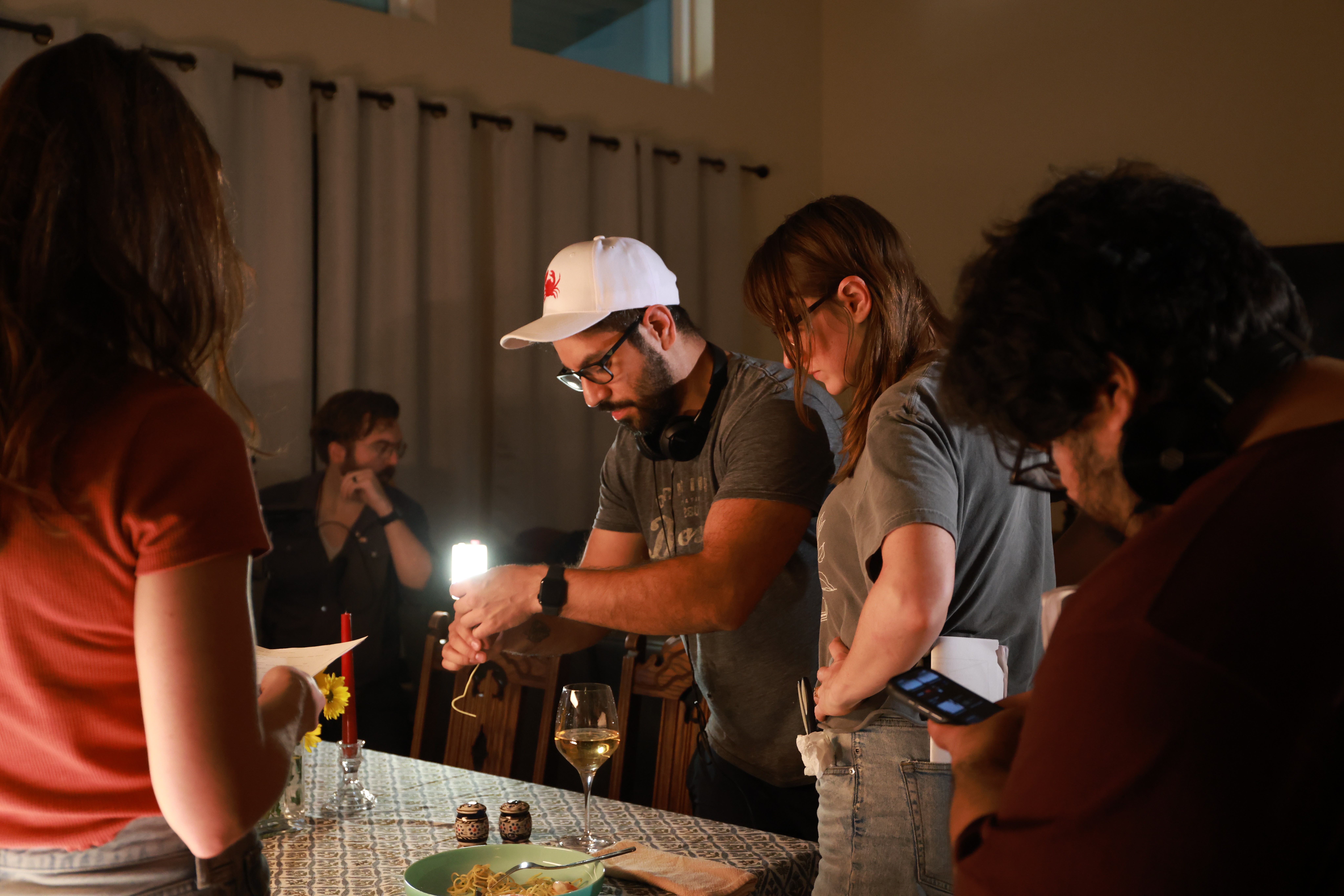 Group of people gathered around a table, some using phones, in a room with curtains and warm lighting.