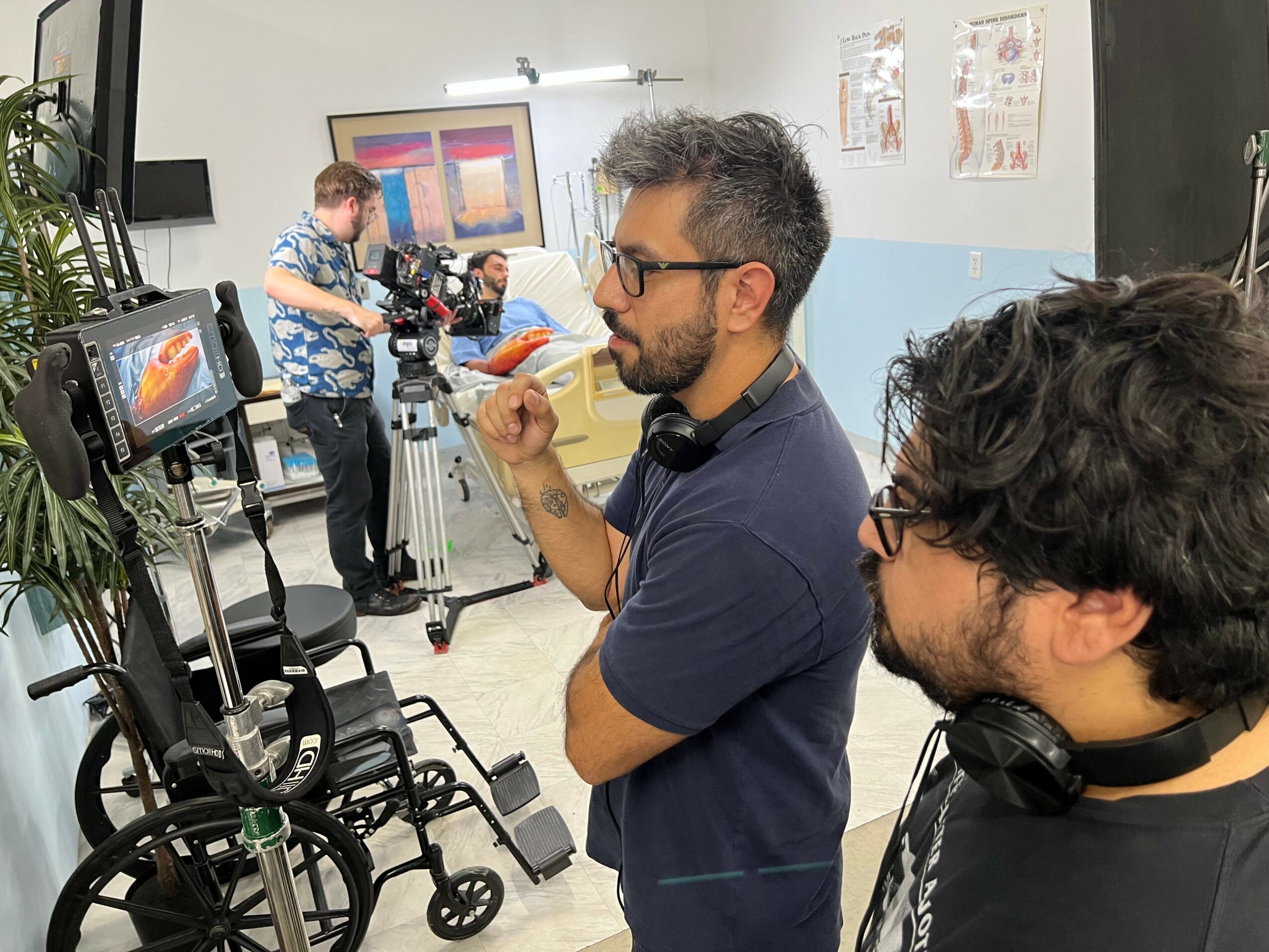 Two men with headphones and glasses in a room with cameras and a person in the background, possibly filming or photographing.