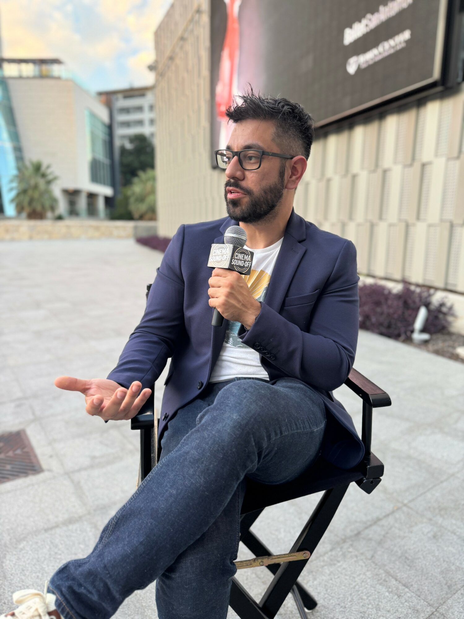 Man with glasses and beard sitting outdoors, holding a microphone, wearing a blazer and jeans, gesturing with his hand.
