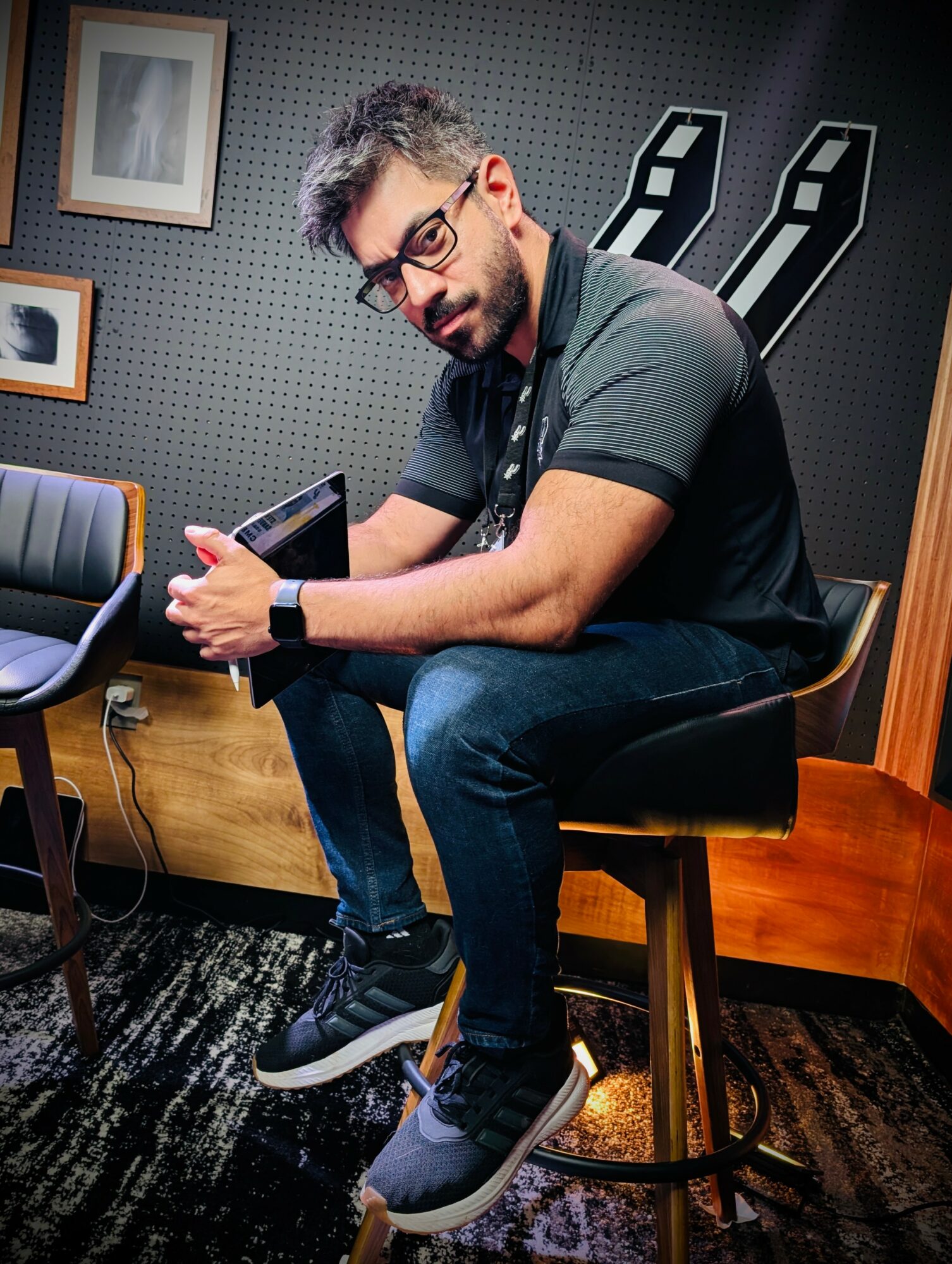 Man sitting on a stool, looking at his phone, in a room with dark walls and framed pictures.