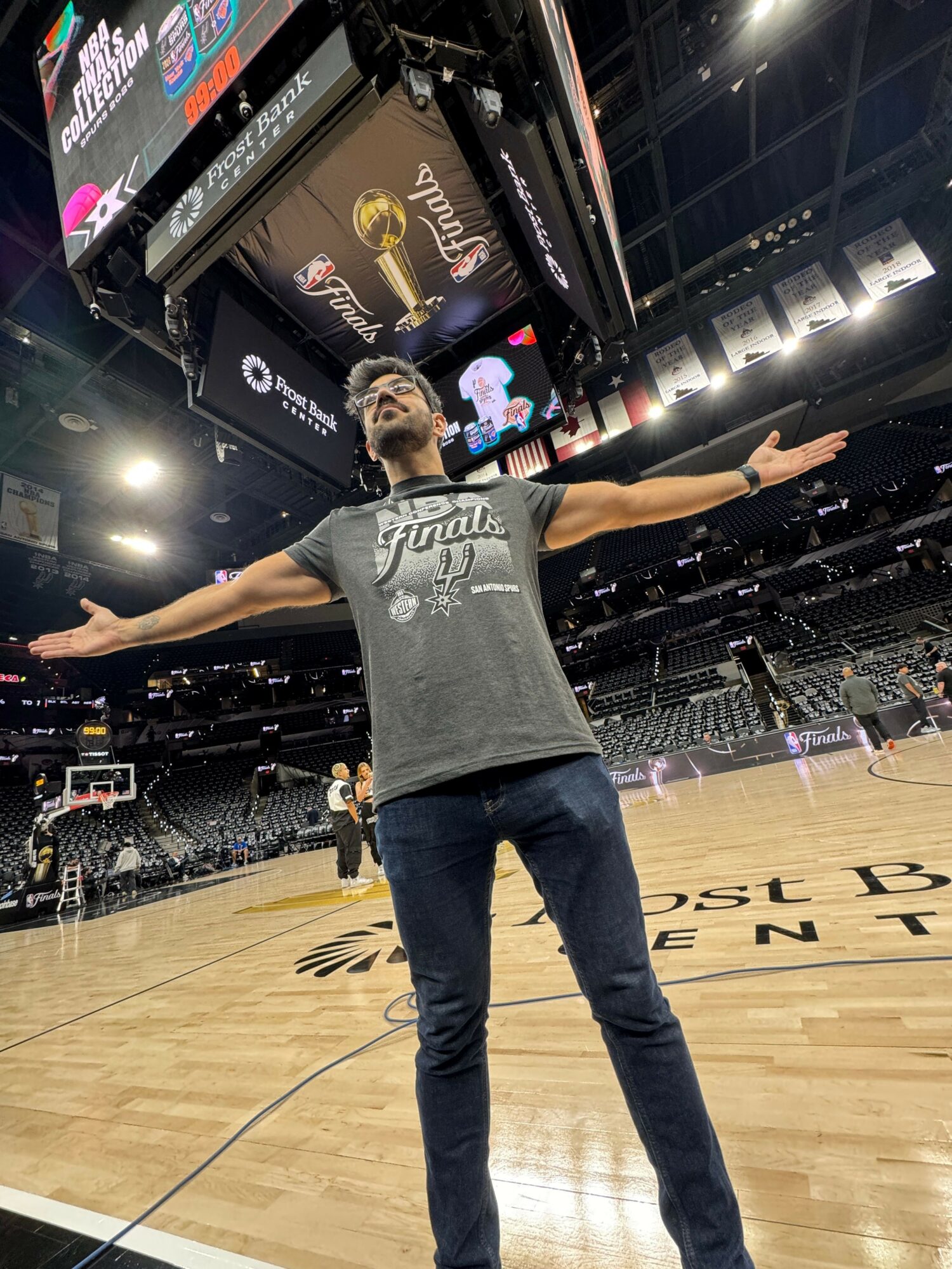 Person standing on a basketball court with arms outstretched, facing a scoreboard and electronic displays, in an indoor arena.