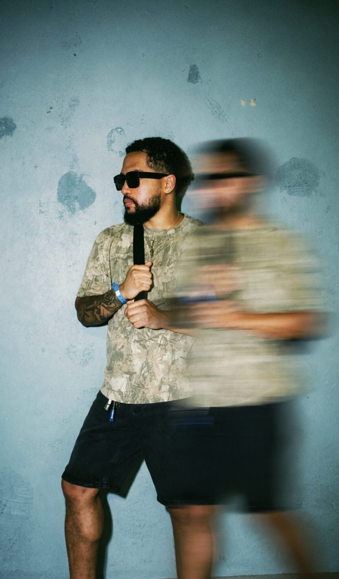 Man with sunglasses holding a microphone, standing against a textured blue wall, with a blurred person nearby.