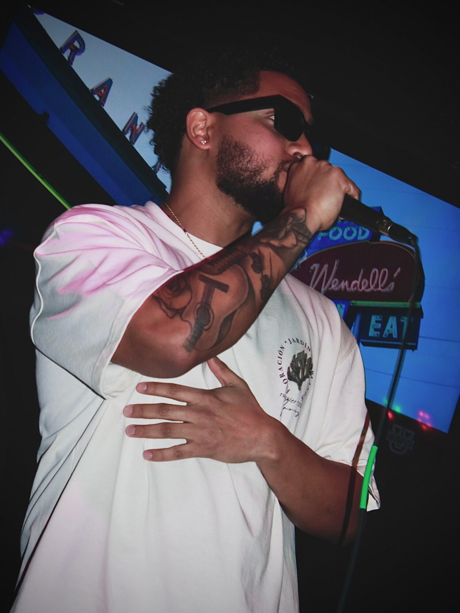 Person with sunglasses and tattoos singing into a microphone, holding their chest, in a dark setting with colorful background lights.