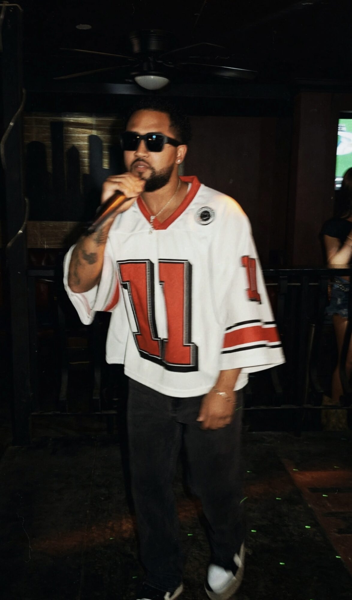 Man with sunglasses and beard, wearing a white sports jersey with red and black details, standing indoors in a dimly lit setting.