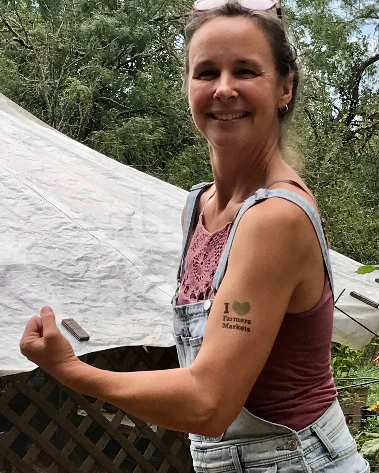 Smiling woman with a tattoo on her upper arm, outdoors with trees and a tent in the background.