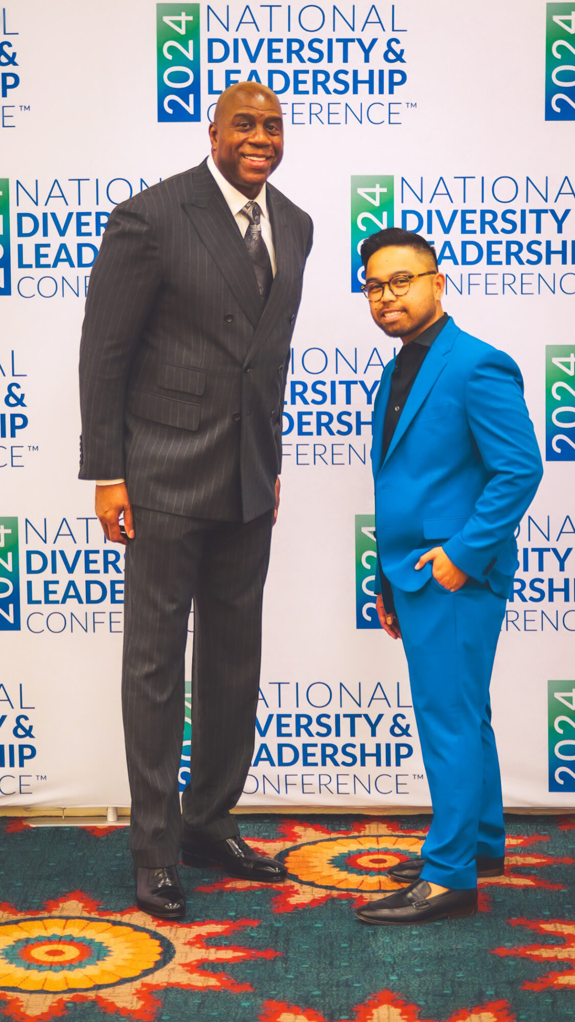 Two men in suits standing on a patterned carpet in front of a backdrop with text about diversity and leadership conference.