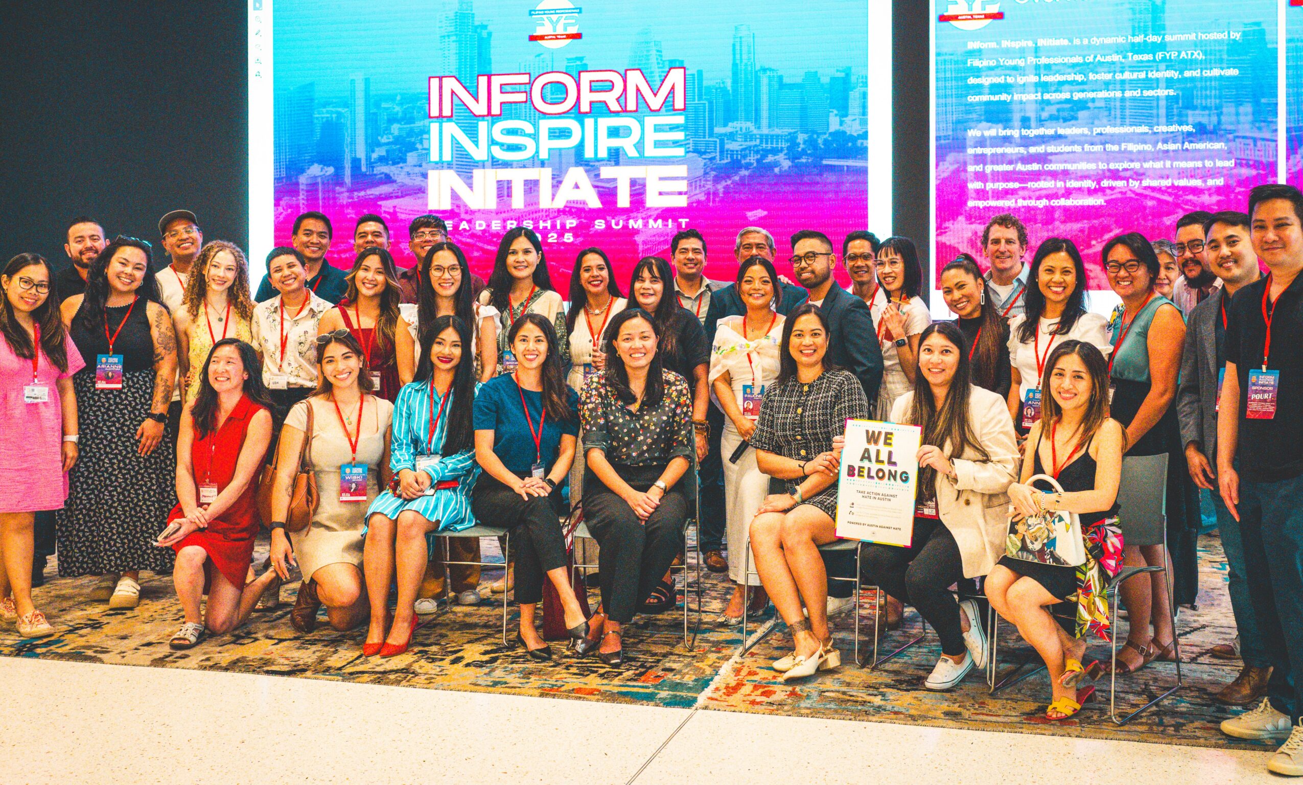 Group of people posing in front of a colorful digital display with text, some holding signs, indoors.