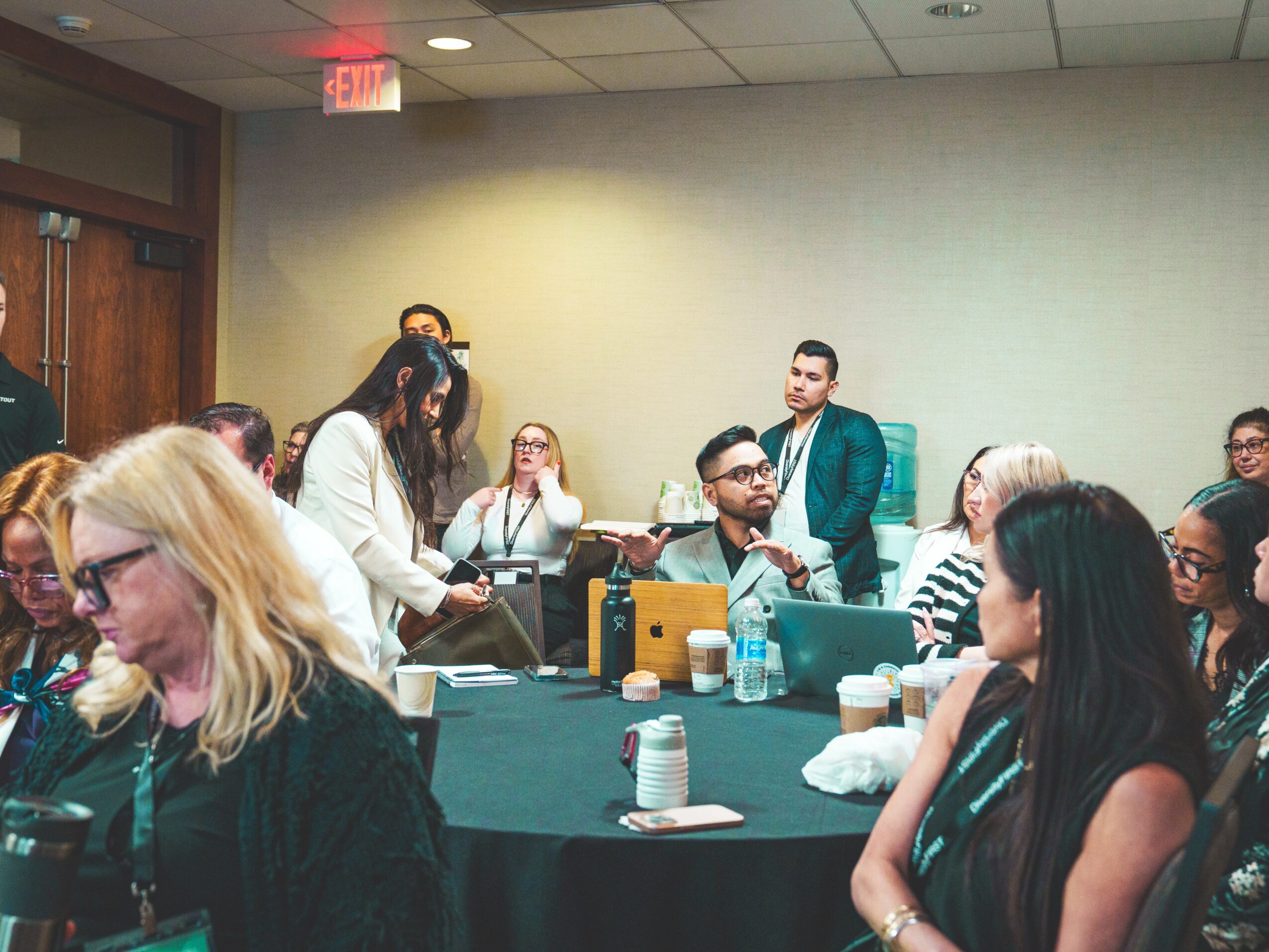 Group of people in a meeting room, some seated and some standing, with tables, papers, and water bottles present.