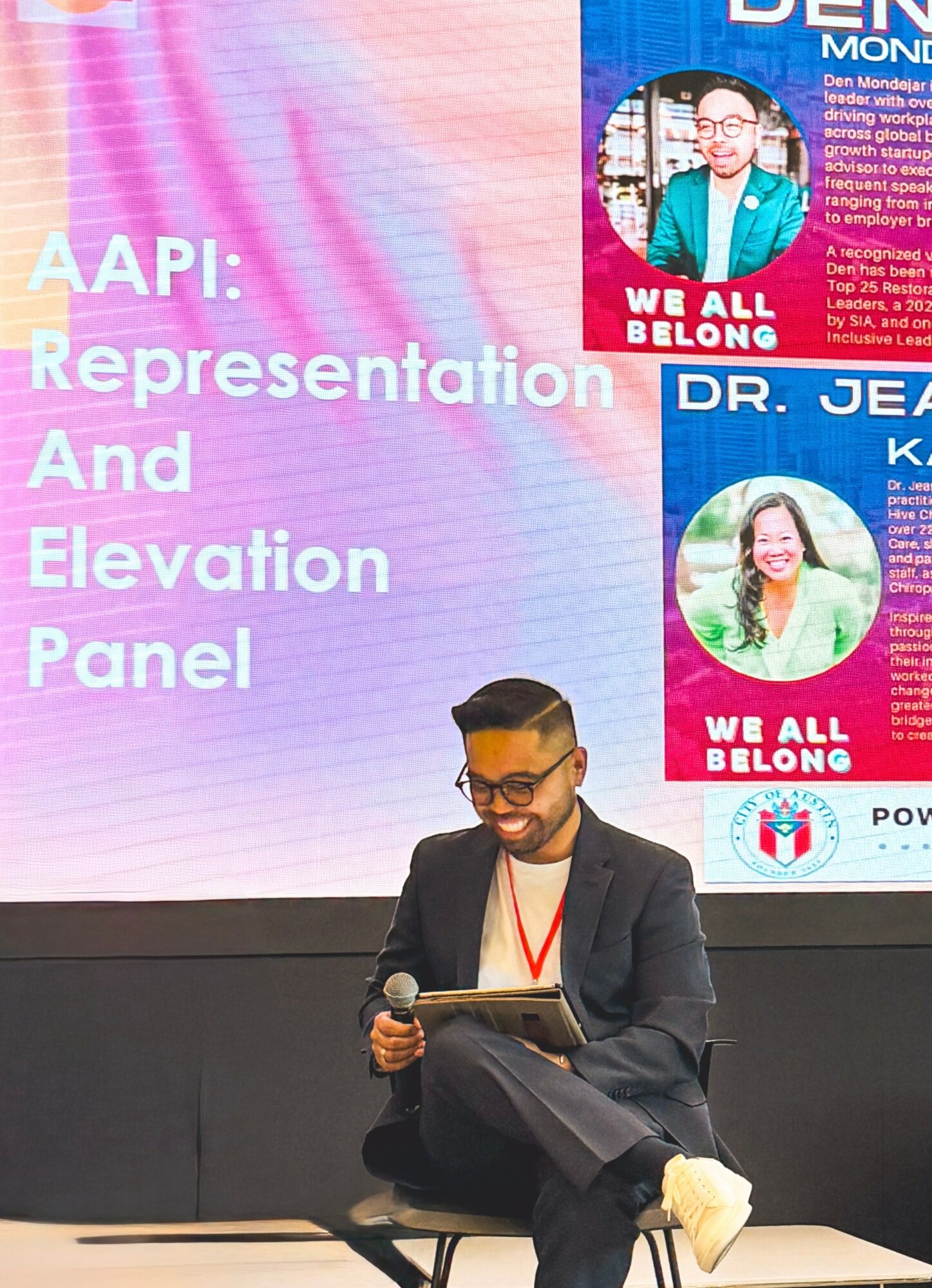 Person sitting on stage with a microphone, wearing glasses and a suit, in front of a presentation screen with text and images.