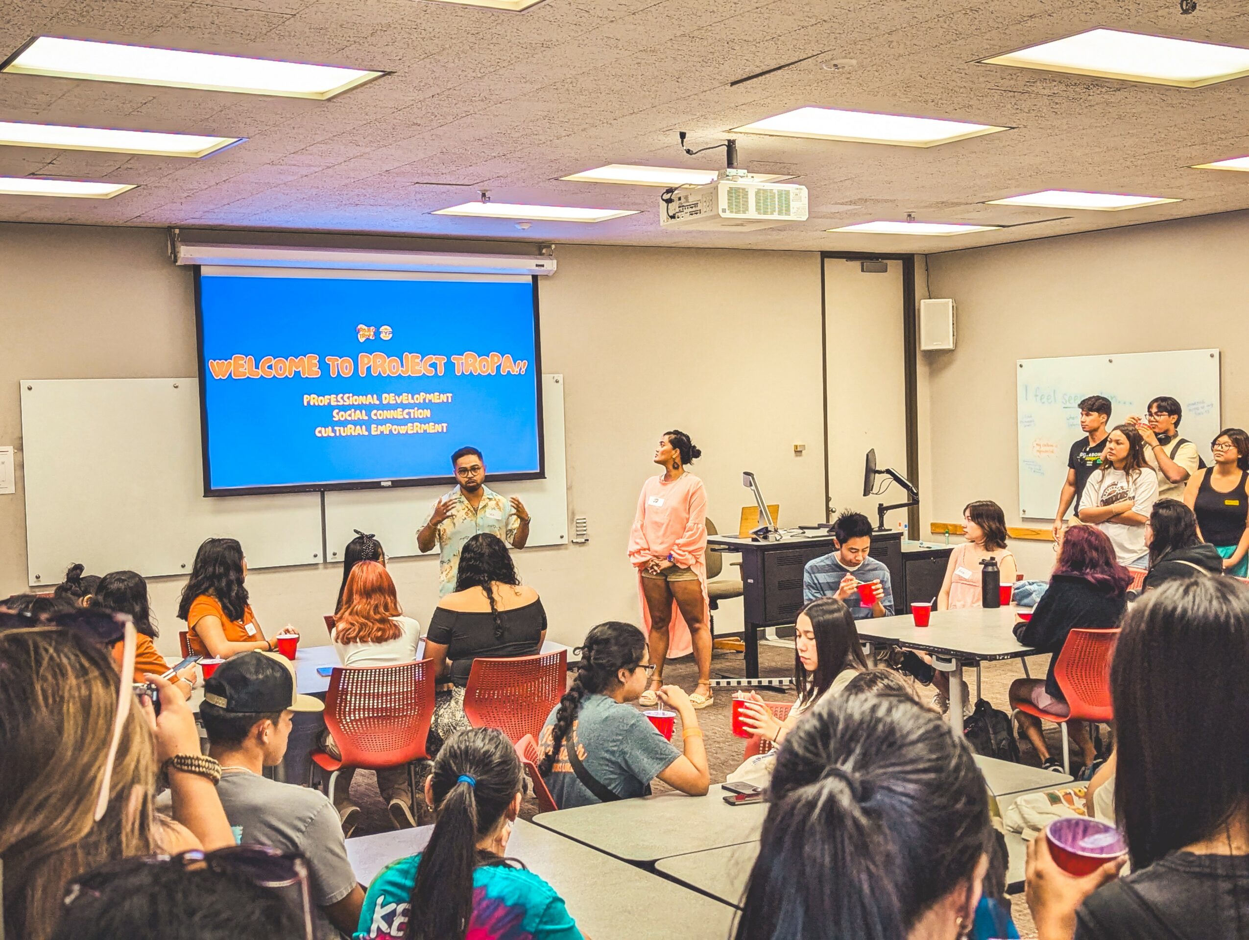 Classroom with students and teachers, presentation screen displaying welcome message, and people standing at the front.