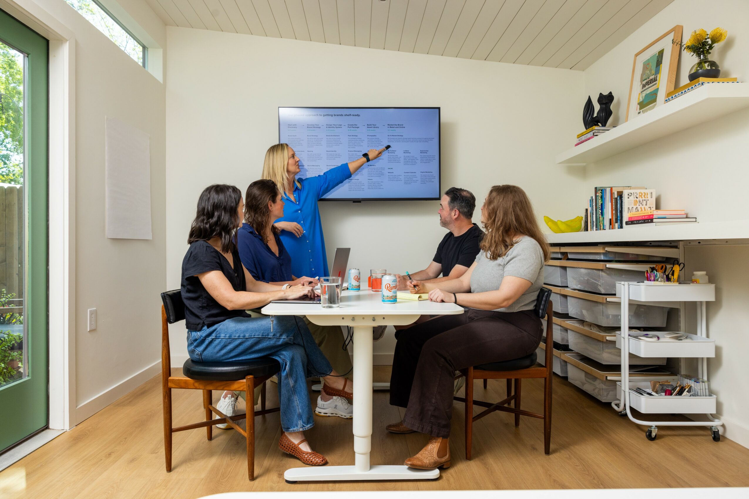 Five people in a meeting room, one woman standing and pointing at a screen, others seated around a table, books and shelves visible.