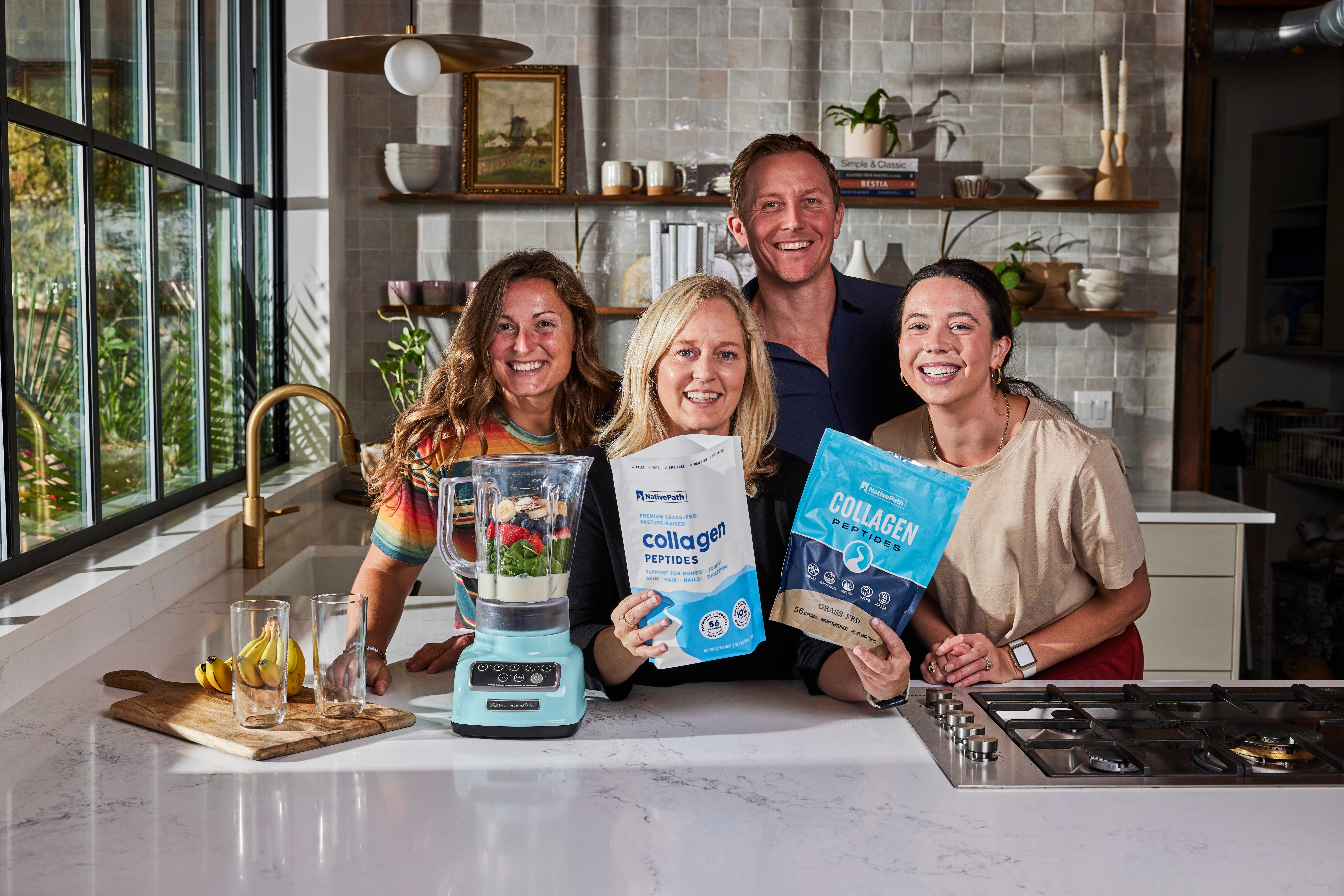 Four smiling people in a kitchen holding collagen supplement packages, standing behind a white counter with a blender and food items.
