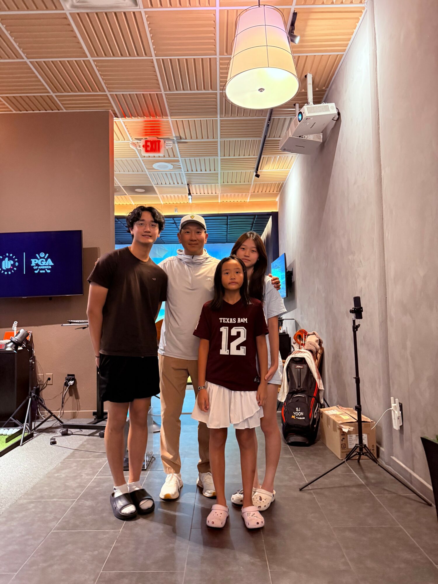 Four people standing indoors, with a ceiling light, wall, and equipment in the background.