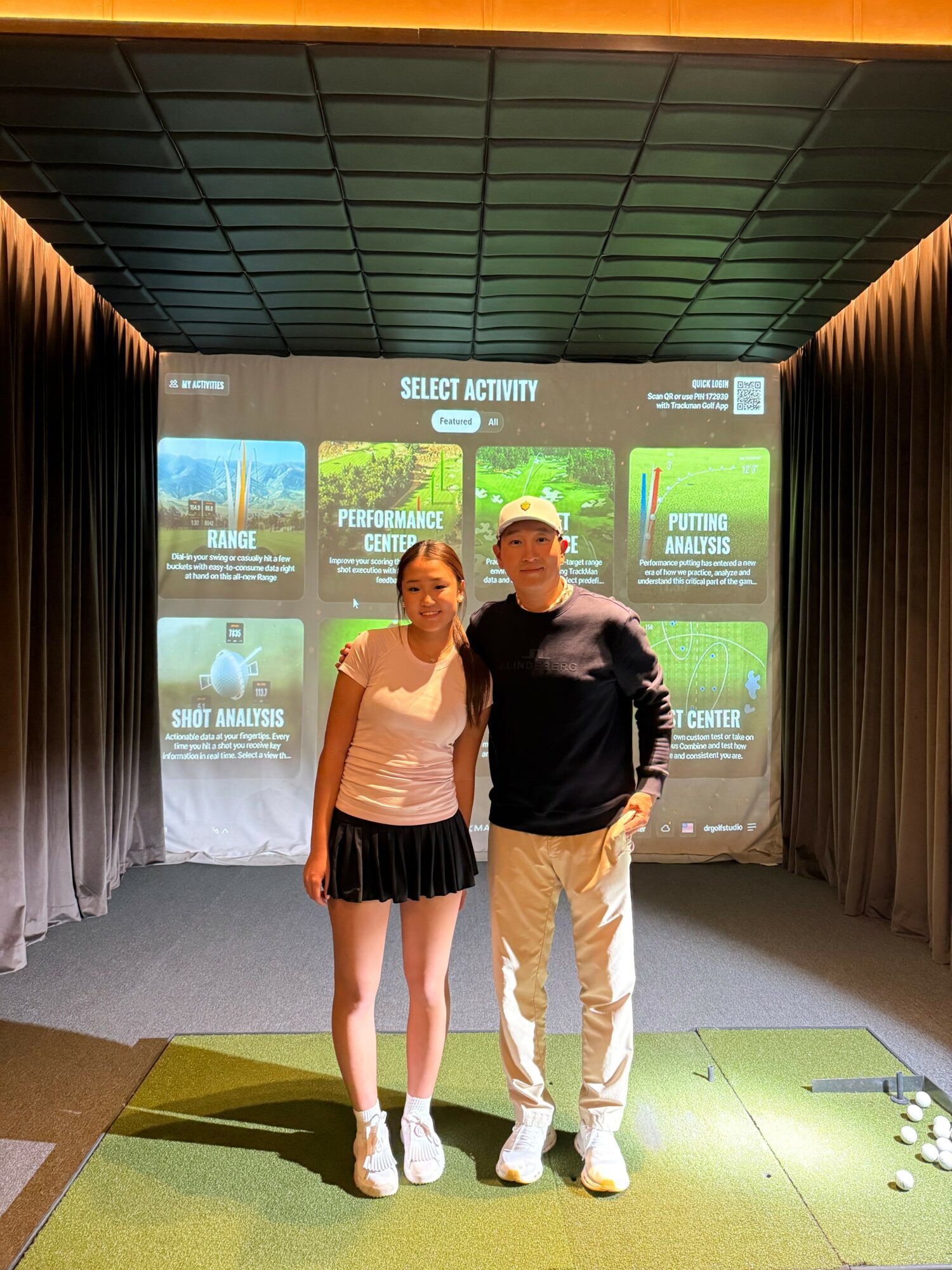 Two people standing on a stage in front of a large screen displaying activity options, with curtains on sides and a green carpeted floor.