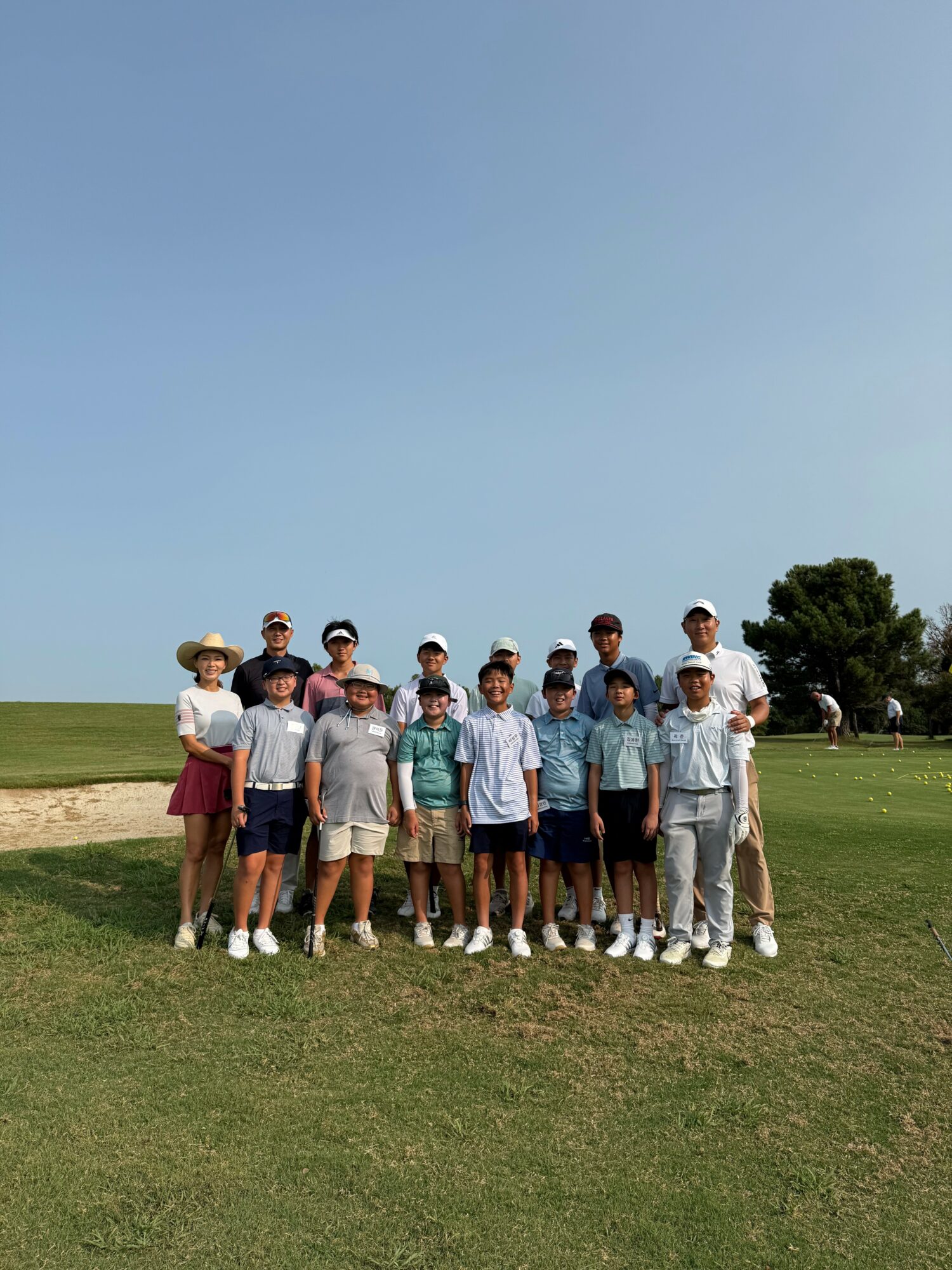 Group of people standing outdoors on grass under a clear blue sky, some wearing hats and casual clothing.