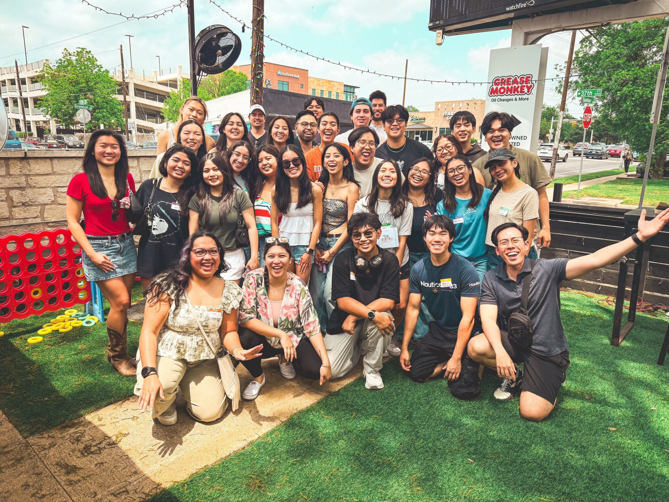 Group of 24 people outdoors on grass, smiling, posing for photo, some making gestures, background includes buildings, trees, and a sign.