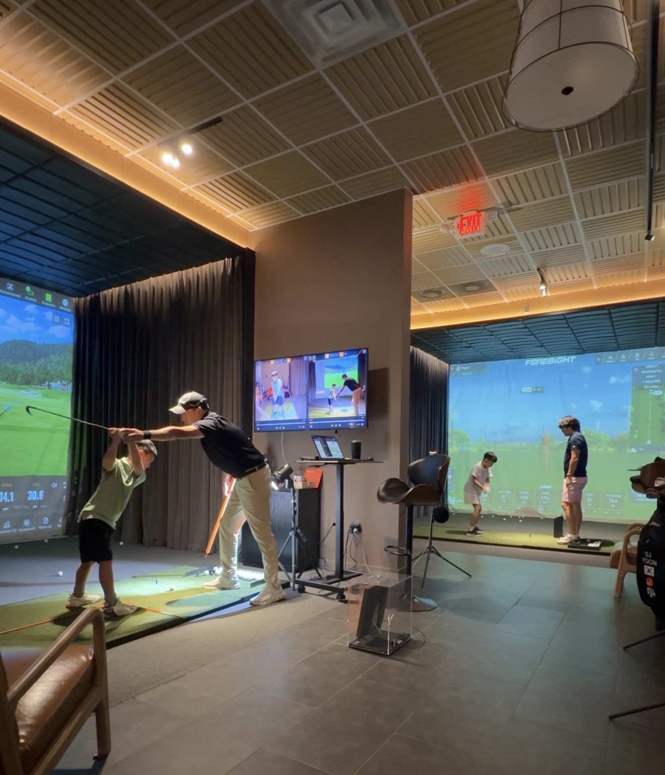 Indoor golf simulation room with children and an adult playing, large screens showing golf courses, and a ceiling with lights.