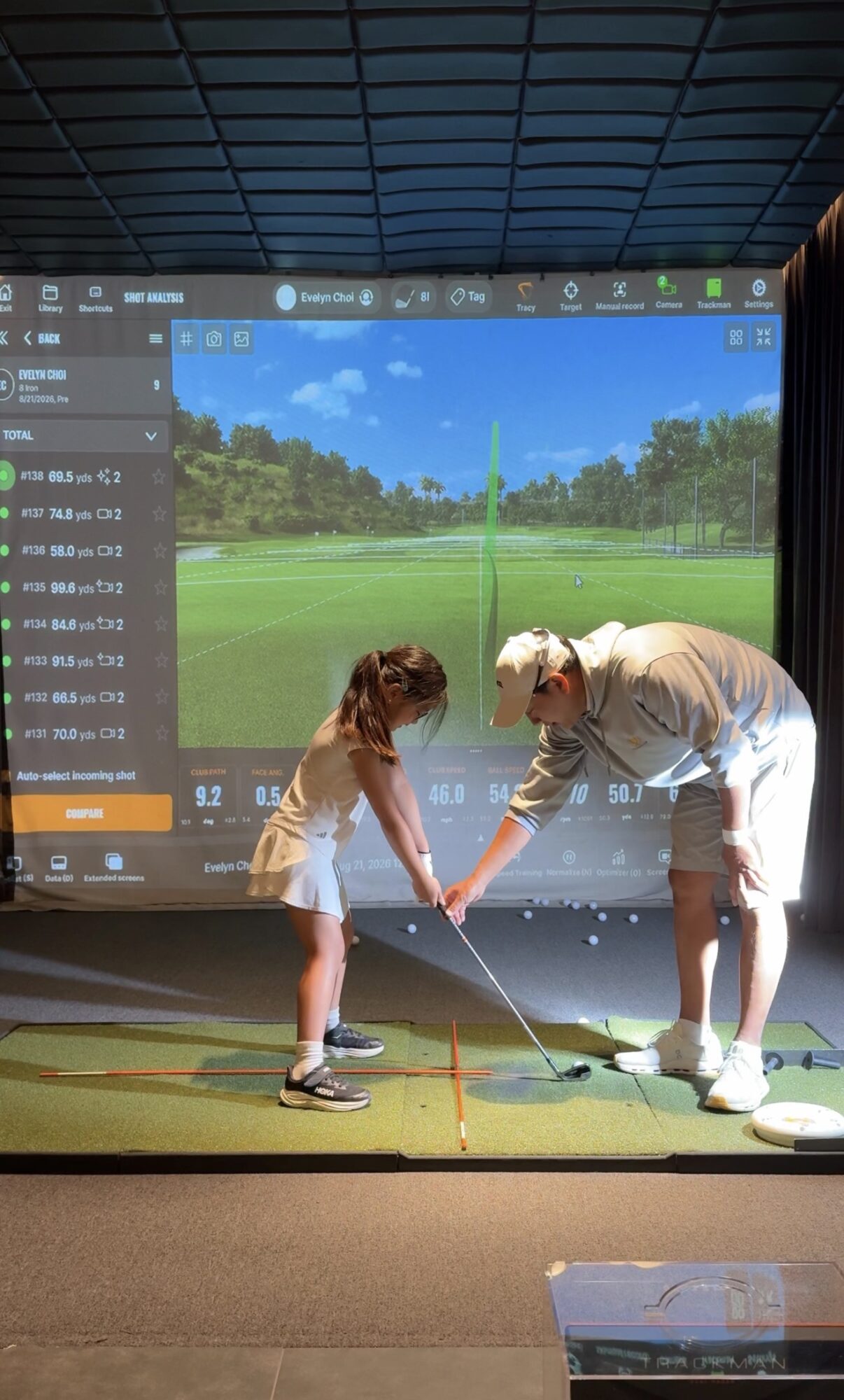 Two children and an adult at an indoor golf simulator, holding a golf club together, with a virtual golf course on the screen behind them.