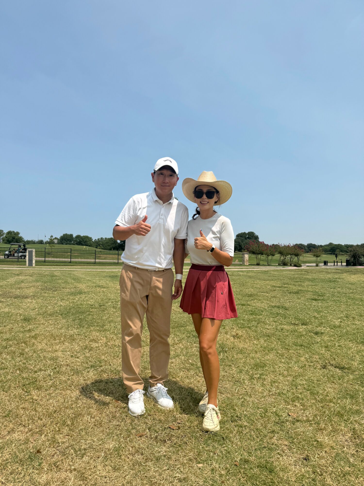 Two people standing on a grassy field under a clear blue sky, one wearing a white cap and the other wearing a wide-brimmed hat and sunglasses, both smiling and posing for the photo.
