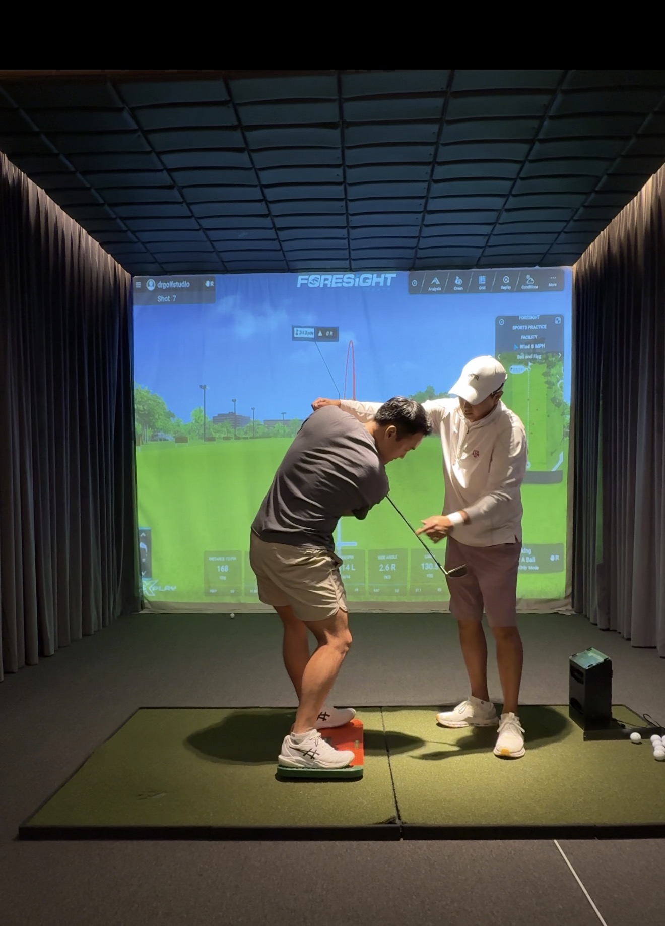 Two people stand on a green mat in front of a large screen displaying a golf simulation, one person teaching the other.
