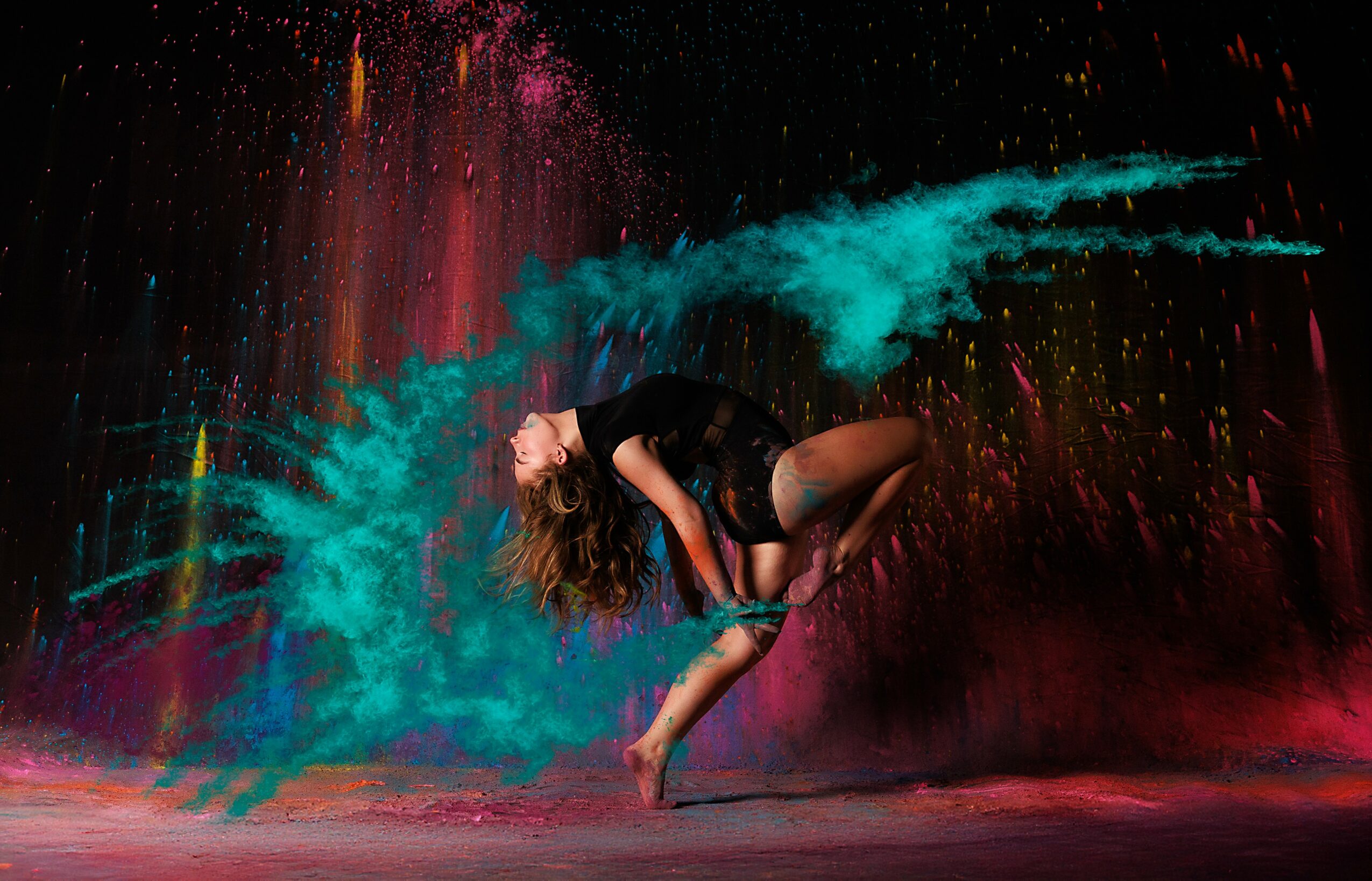 Person performing dance move with colorful powder explosion background, mid-air pose, dark clothing, and vibrant colors.