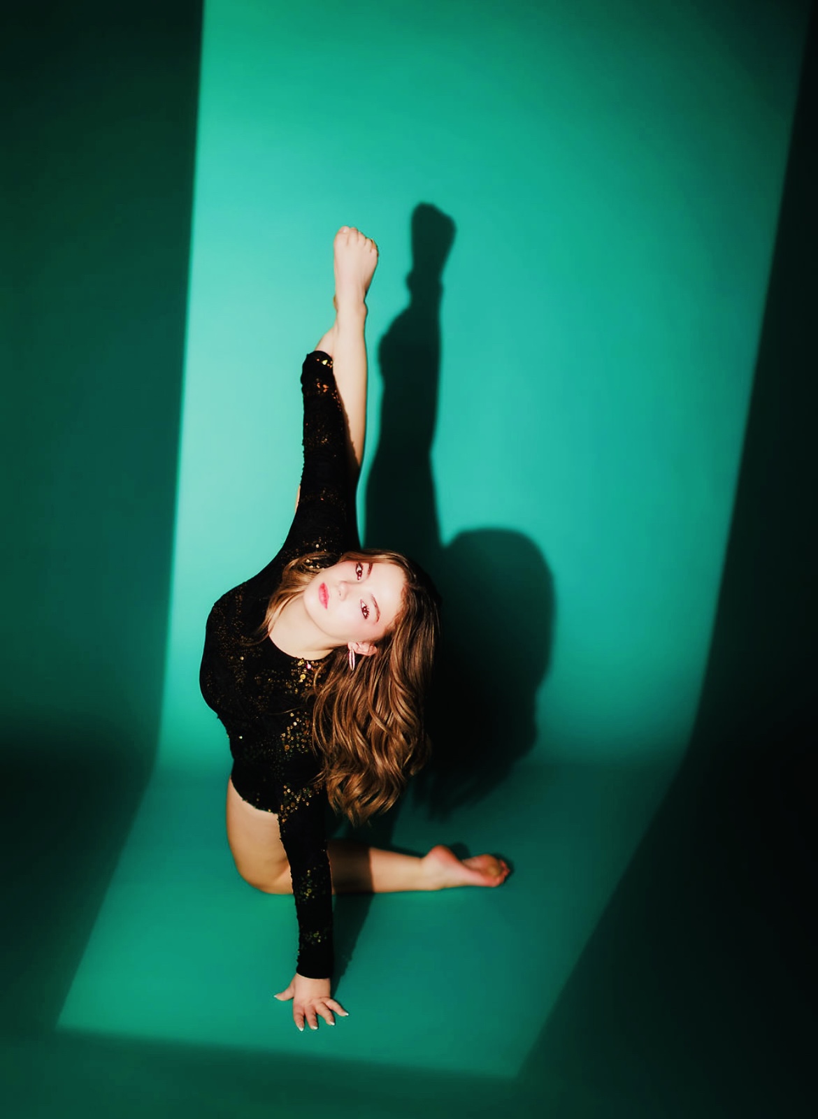 Person performing a contortion pose against a green background, with one leg extended upward and head tilted sideways.