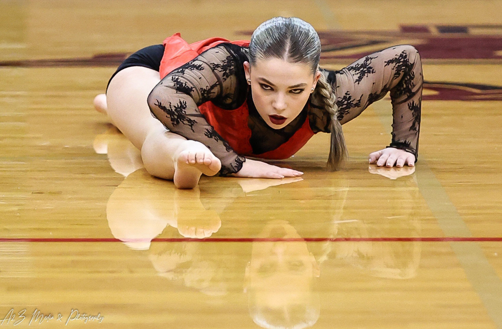 A female gymnast is on the floor in a gymnasium, wearing a black and red leotard with lace sleeves, looking forward.
