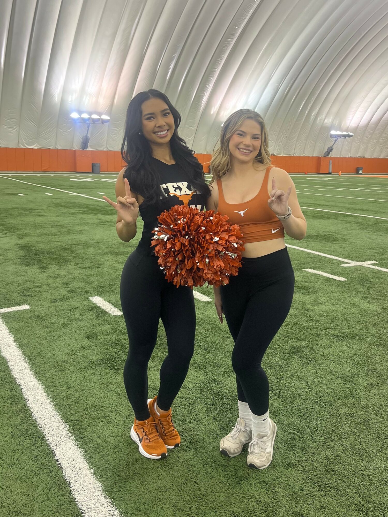 Two women standing on an indoor football field, smiling, holding pom-poms, one making a peace sign, in athletic clothing.