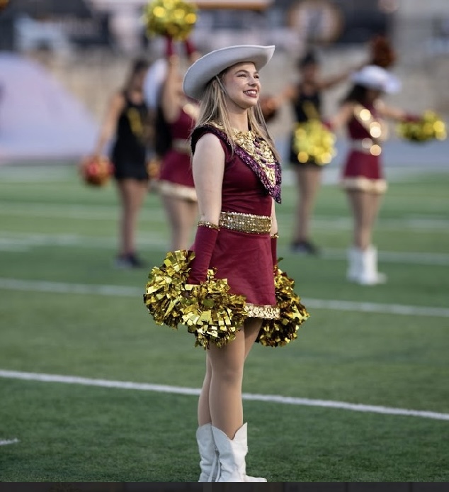 Cheerleader in maroon and gold uniform holding pom-poms on a football field, wearing a white hat and boots, smiling.