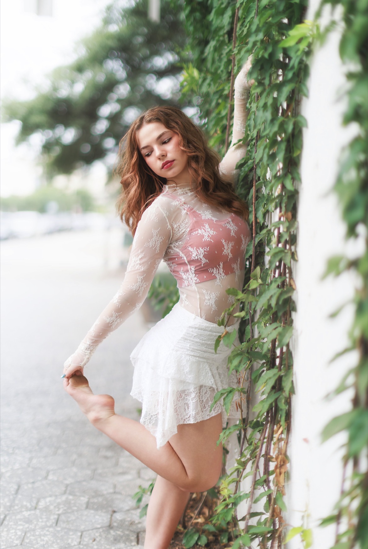 Young woman with curly hair in a pink lace top and white skirt standing next to a vine-covered wall outdoors.