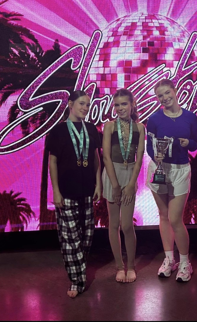 Three young girls stand on stage with medals and a trophy, in front of a pink and purple backdrop with a disco ball.