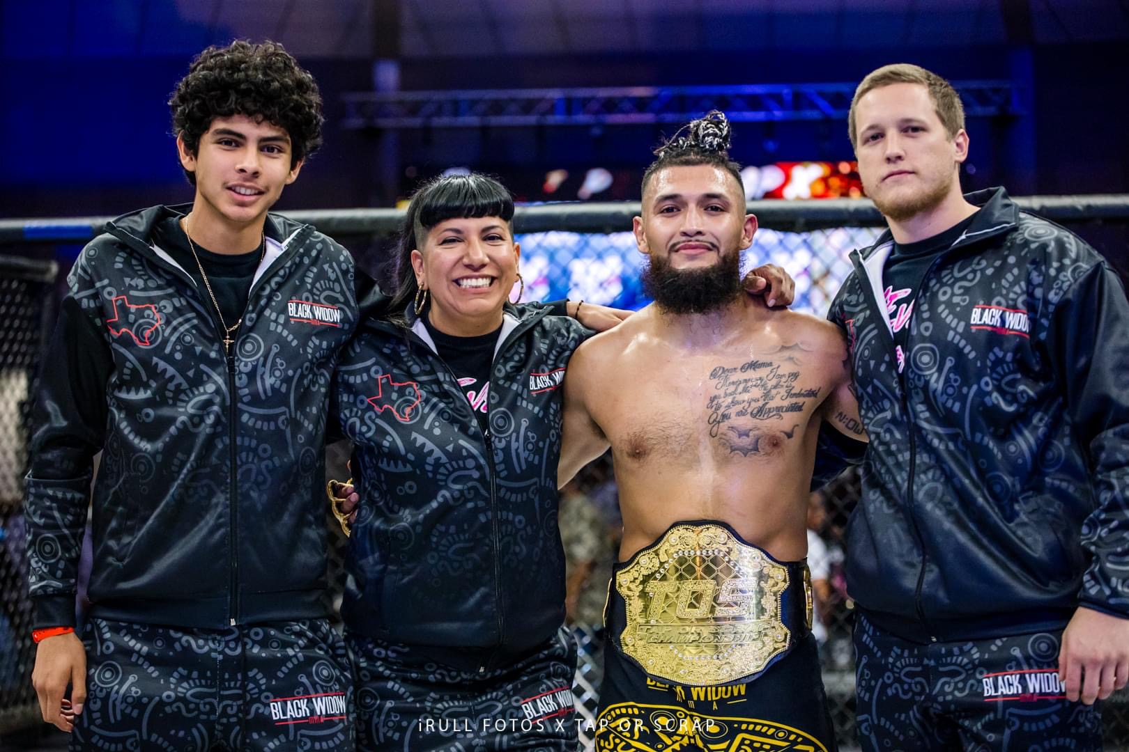 Four people, one shirtless with a championship belt, standing together in a sports arena, smiling at the camera.
