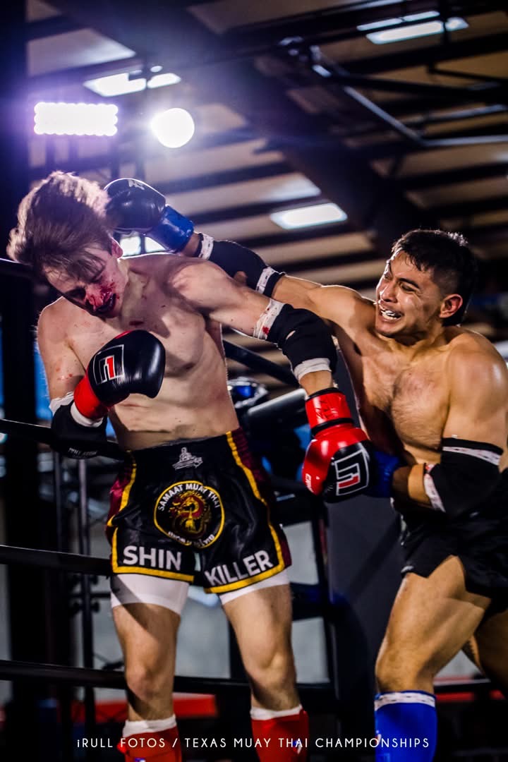 Two male Muay Thai fighters in a ring, one delivering a punch to the other's face, both wearing gloves and shorts.