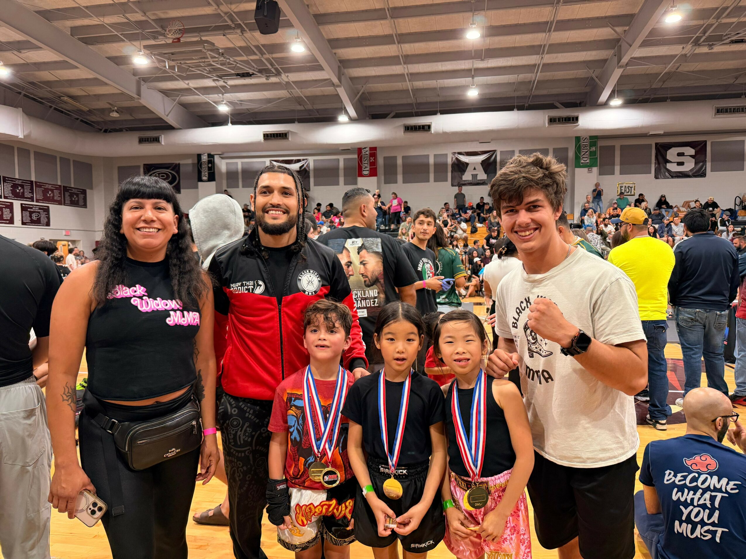 Group of five people, including children with medals, in a gymnasium with many spectators and banners in the background.