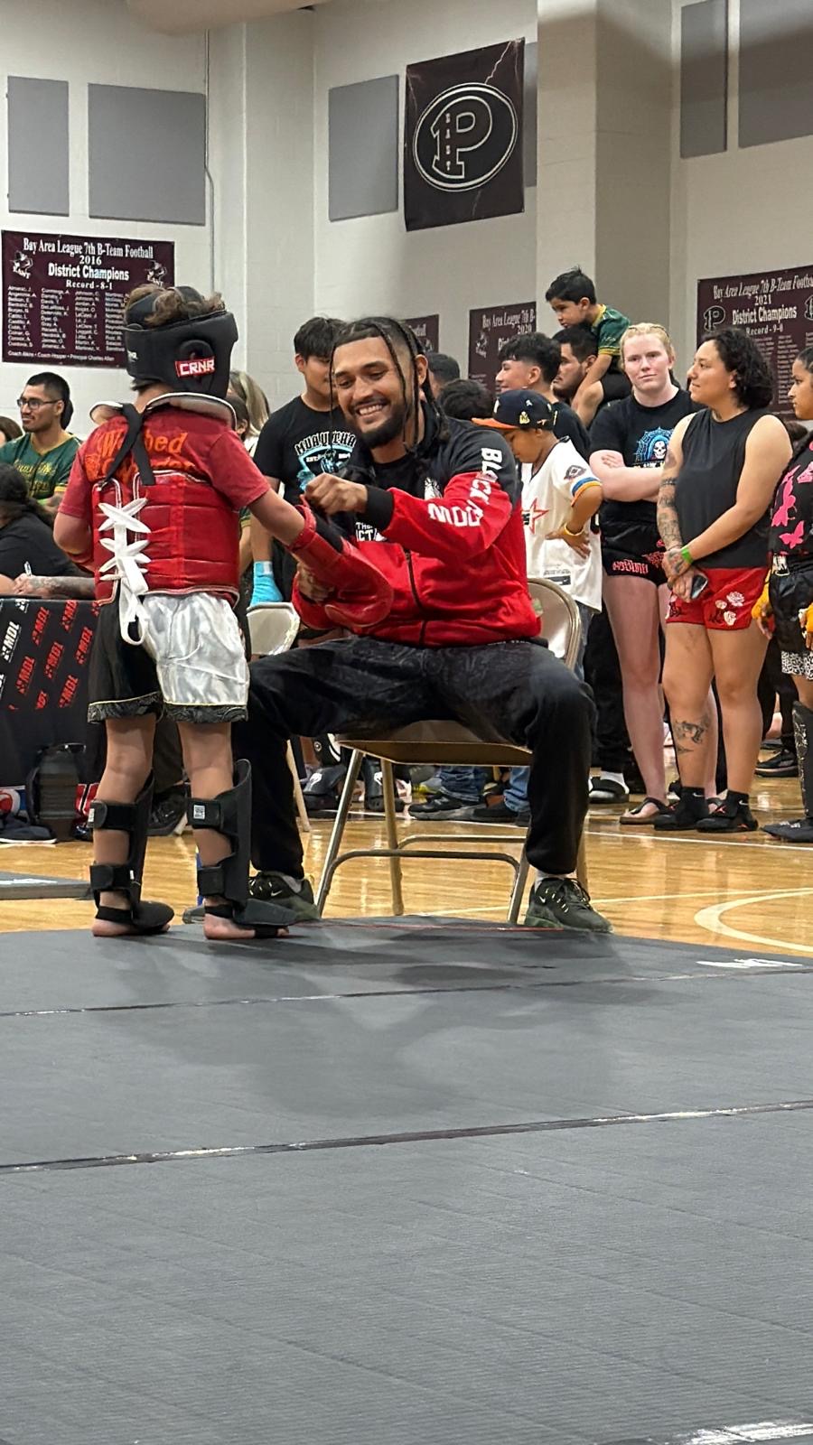 Man sitting on a chair smiling, surrounded by children and adults in a gymnasium, some in costumes, with a crowd in the background.
