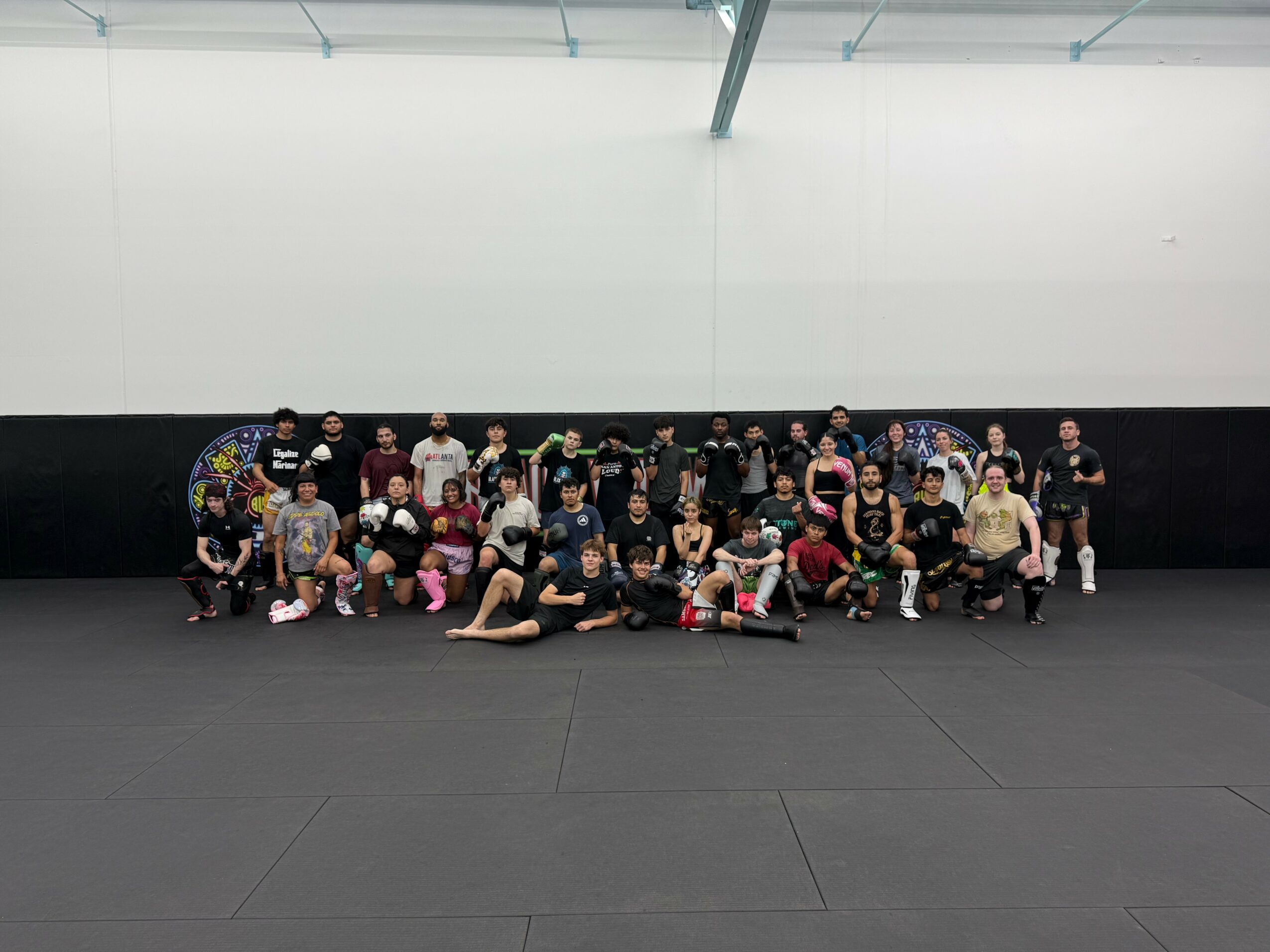 Group of children and adults posing in a gym, some sitting and some standing, with a white and black wall background.