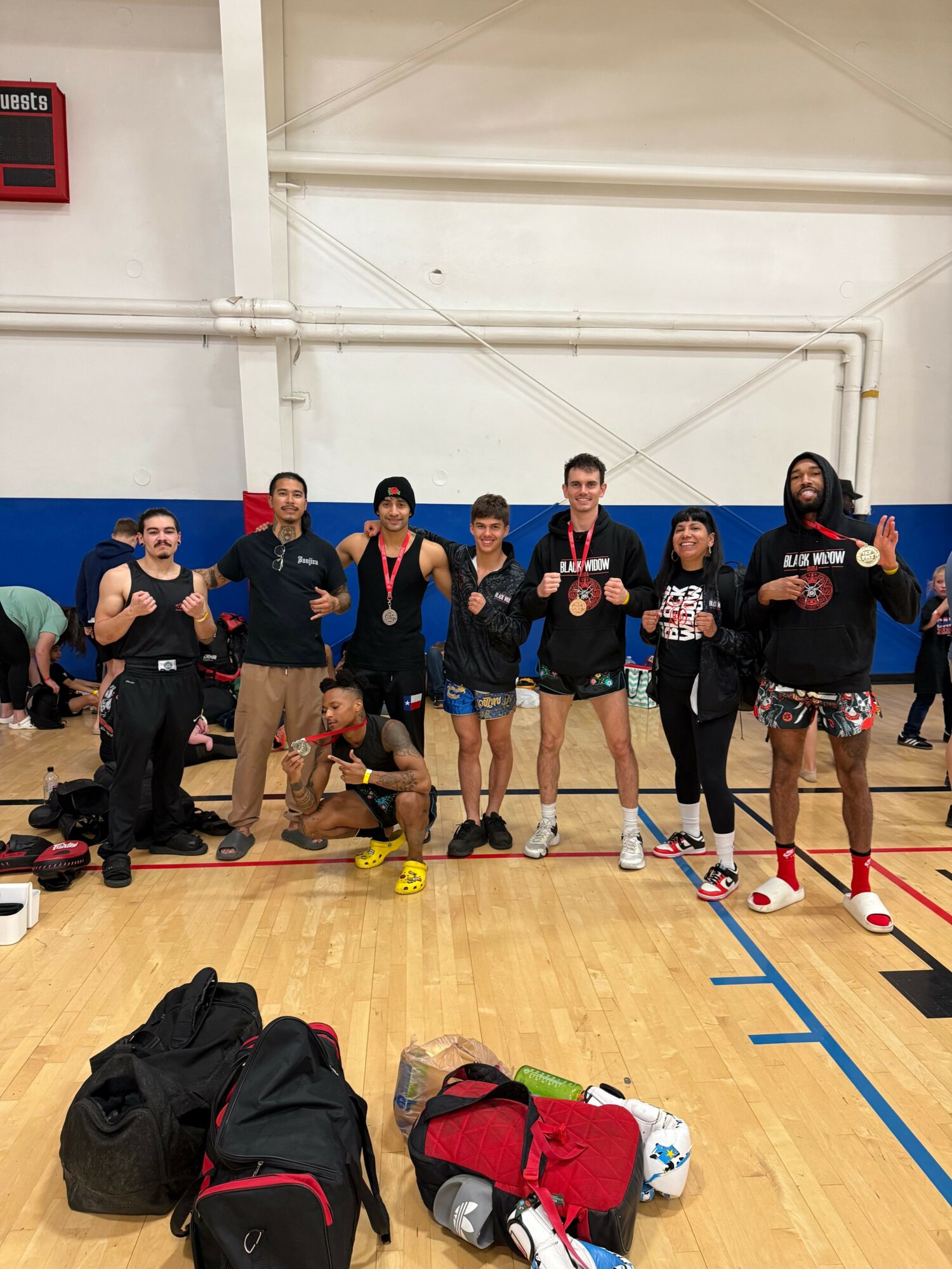 Group of people in sportswear standing on a gymnasium floor, some holding medals, with bags and sports equipment nearby.