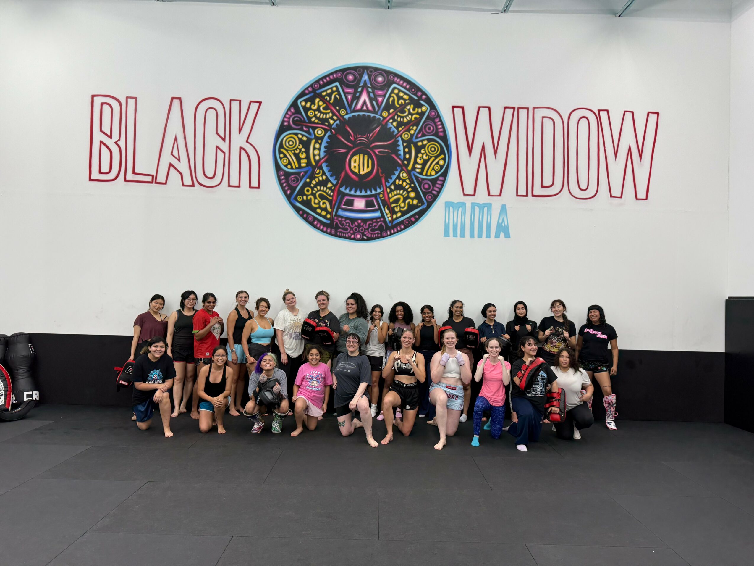 Group of people posing in front of a wall with a colorful circular design and the words 'Black Widow MMA'.